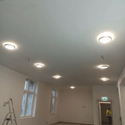 Paweł Herasimov - Wnętrze po montażu oświetlenia: okrągłe lampy sufitowe równomiernie rozmieszczone w przestronnym pomieszczeniu z oknami i drabiną malarską.