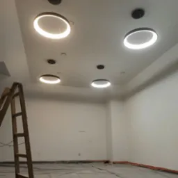 Nowoczesne, okrągłe lampy LED zamontowane na suficie w pustym pomieszczeniu. Widoczna drabina, zabezpieczona podłoga folią malarską.