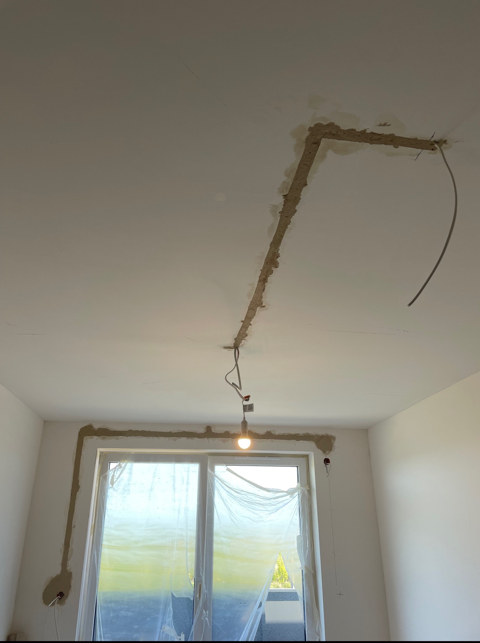 Wykute bruzdy pod kable elektryczne wzdłuż sufitu i ściany nad oknem, zabezpieczonym folią. Widoczna żarówka na kablu. Etap prac przed tynkowaniem.