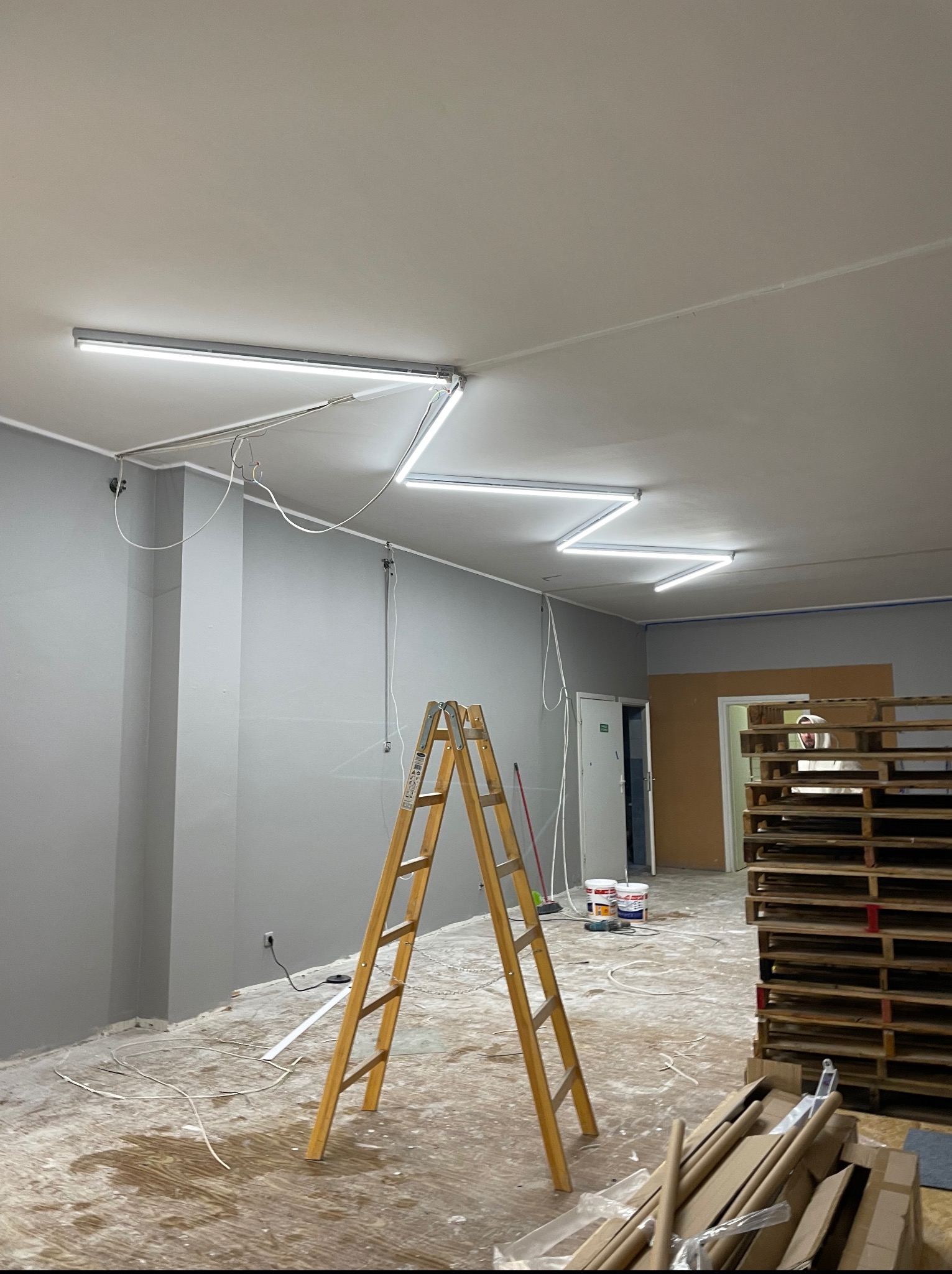 Montaż oświetlenia LED w pomieszczeniu z drabiną i paletami. Widoczne przewody i puszki instalacyjne. Szare ściany i sufit. Wnętrze w trakcie wykończenia.