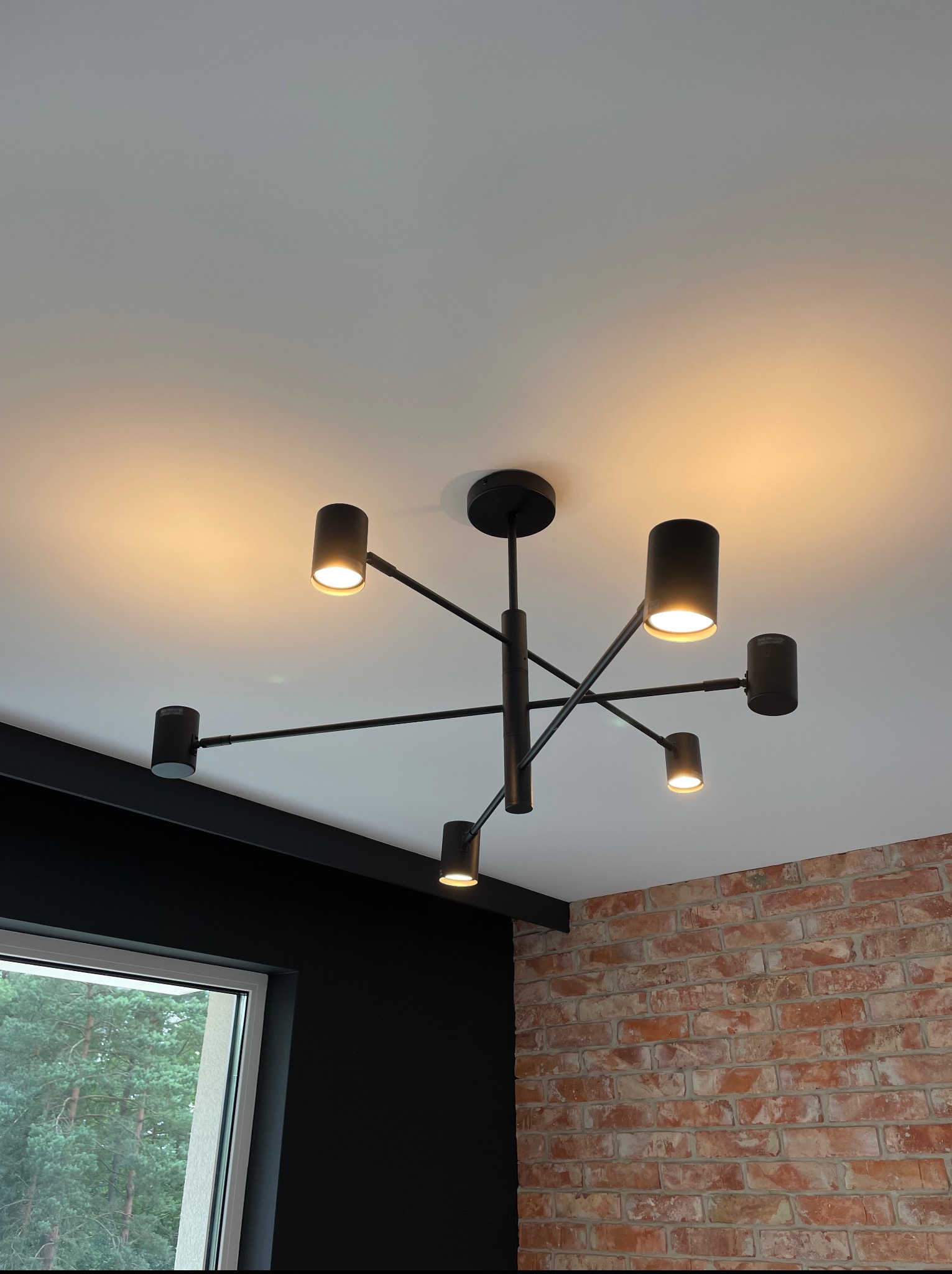 Czarny, nowoczesny żyrandol sufitowy z sześcioma punktami świetlnymi, zamontowany w pomieszczeniu z ceglaną ścianą i czarną ramą okienną.