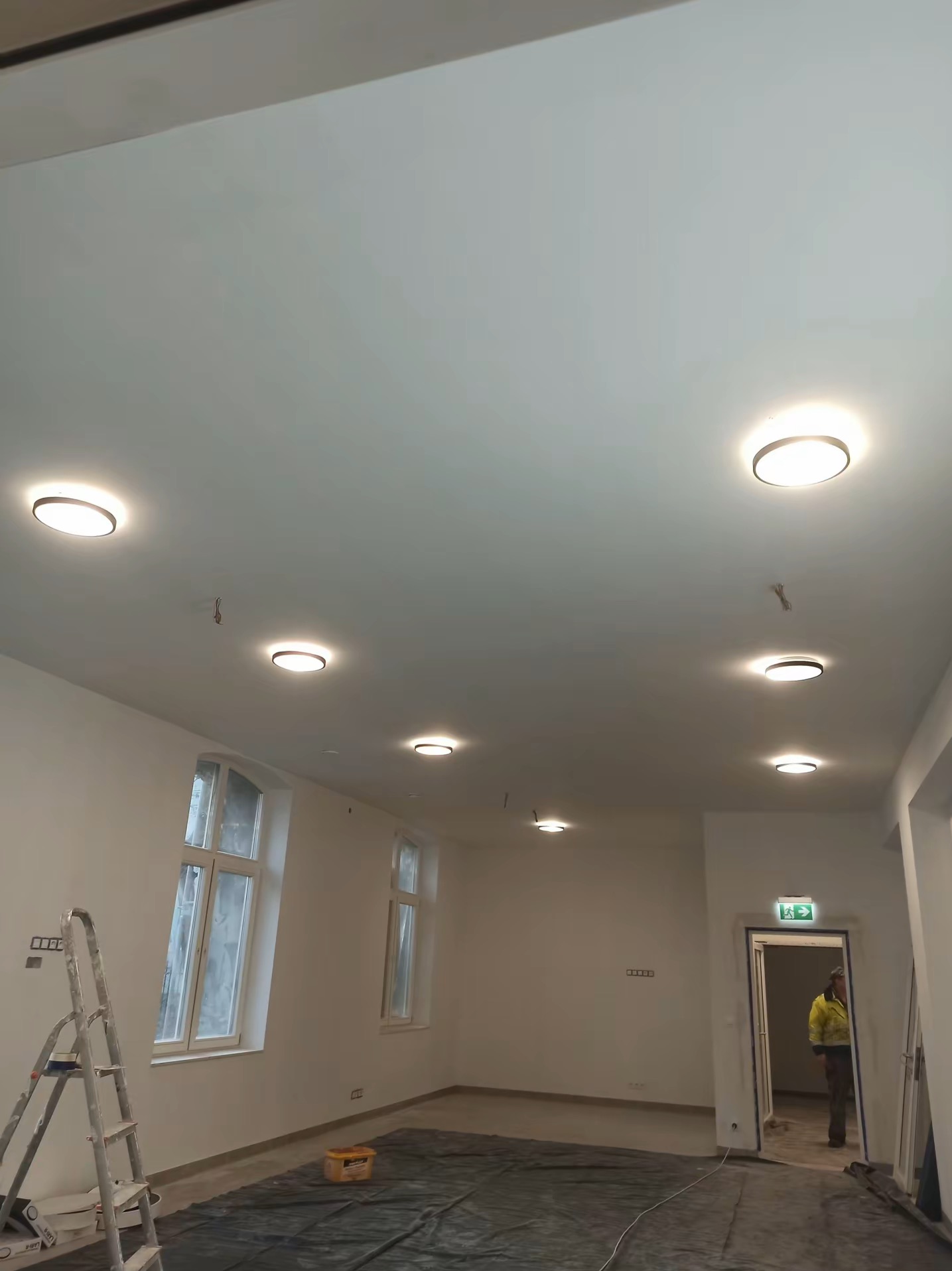 Wnętrze po montażu oświetlenia: okrągłe lampy sufitowe równomiernie rozmieszczone w przestronnym pomieszczeniu z oknami i drabiną malarską.