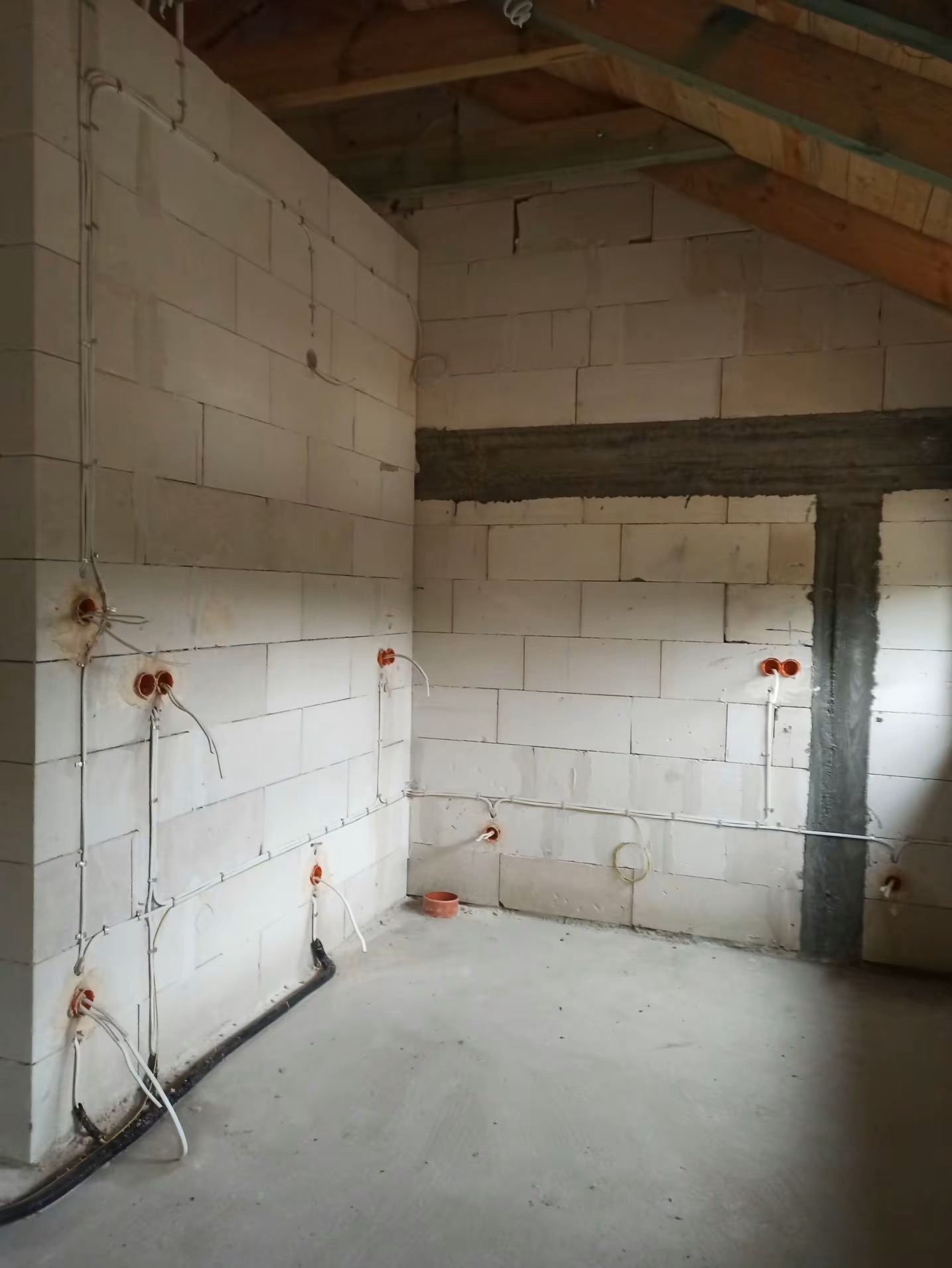 Surowy stan poddasza z instalacją elektryczną: kable prowadzone po ścianach z betonu komórkowego, puszki instalacyjne w kolorze pomarańczowym, widoczne drewniane elementy konstrukcji dachu.