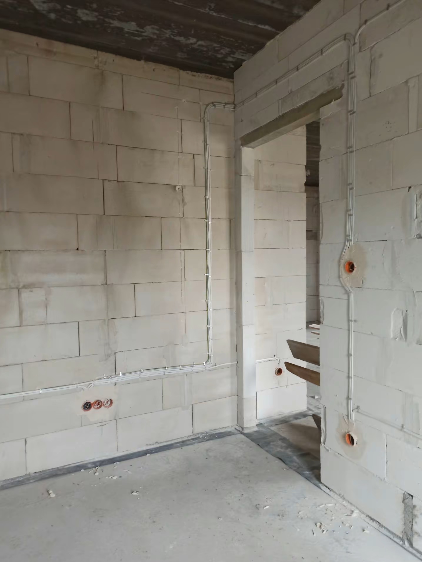 Surowy mur z widocznymi, nowymi instalacjami elektrycznymi w peszlach, gniazda w ścianie, widok na przejście do innego pomieszczenia w stanie deweloperskim.