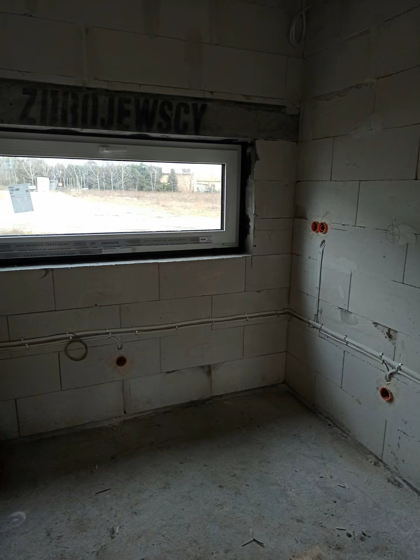 Wnętrze w stanie surowym z widoczną instalacją elektryczną: kable prowadzone po ścianach z betonu komórkowego, puszki elektryczne. Okno z widokiem na zewnątrz.