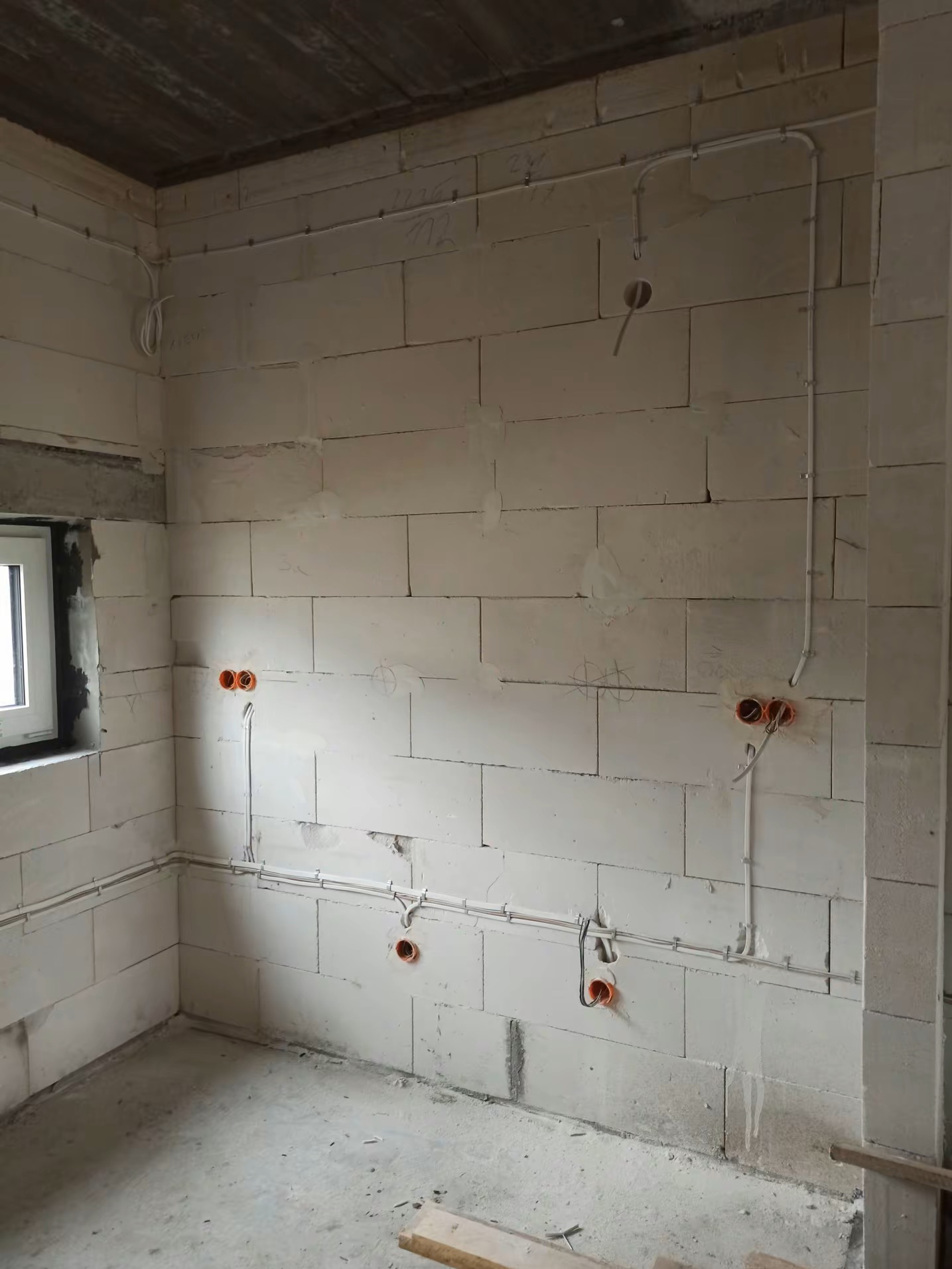 Surowe wnętrze w trakcie prac elektrycznych: widoczne kable w białych listwach, pomarańczowe puszki pod gniazdka na ścianie z białych bloczków, surowy betonowy strop.