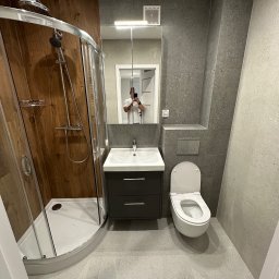 SD Project Sp. z o.o. - Nowoczesna łazienka z kabiną prysznicową z drewnianym wykończeniem, umywalką z szafką i podwieszaną toaletą. Szare płytki na ścianach i podłodze.