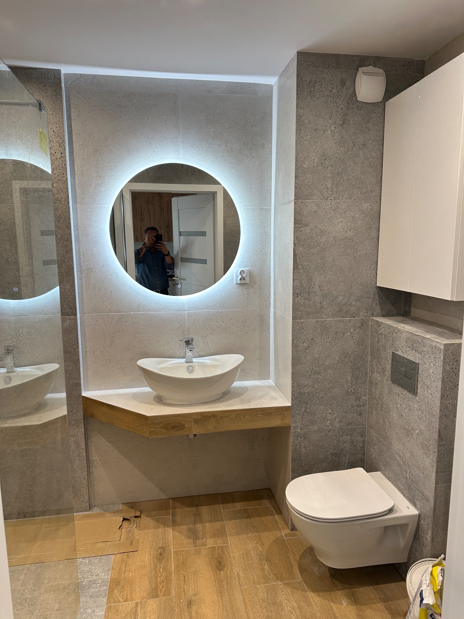 Nowoczesna łazienka z okrągłym lustrem LED, umywalką na drewnianym blacie i podwieszaną toaletą. Szare płytki, drewnopodobne panele. W lustrze odbicie osoby.