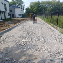 Usługi Transportowo - Budowlane Bartosz Czerwiński - Przygotowanie podłoża pod kostkę: ubijanie kruszywa zagęszczarką. W tle minikoparka i budynki mieszkalne w nowoczesnej architekturze. Krawężniki po bokach.