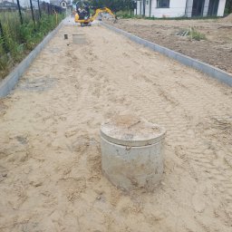 Usługi Transportowo - Budowlane Bartosz Czerwiński - Przygotowanie podłoża pod kostkę: wyrównany piasek, obrzeża betonowe, studzienka kanalizacyjna i koparka w tle. Widoczne ślady opon.