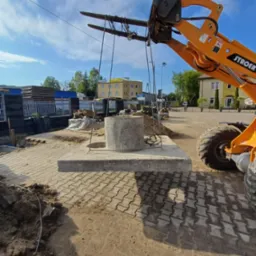 Pomarańczowy wózek widłowy transportuje betonowy element na placu budowy z kostką brukową w tle, widoczny budynek i drzewa.