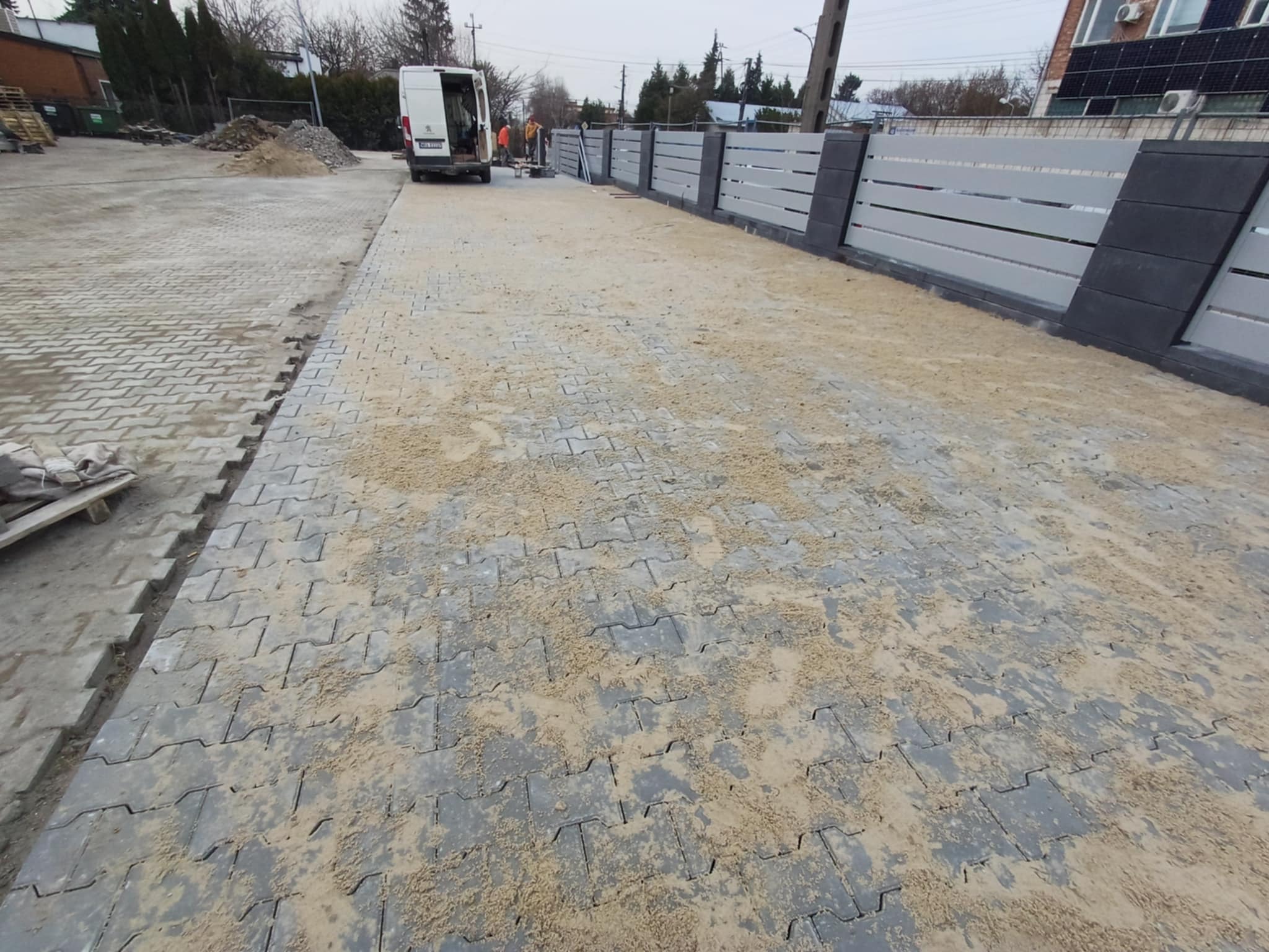 Ułożona szara kostka brukowa na podjeździe, zasypana piaskiem, z widocznym fragmentem ogrodzenia i samochodem dostawczym w tle. Widok z perspektywy ziemi.