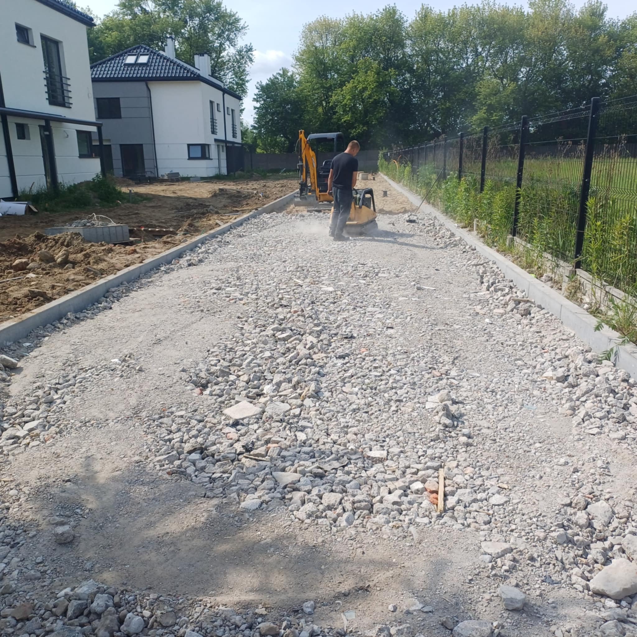 Przygotowanie podłoża pod kostkę: ubijanie kruszywa zagęszczarką. W tle minikoparka i budynki mieszkalne w nowoczesnej architekturze. Krawężniki po bokach.