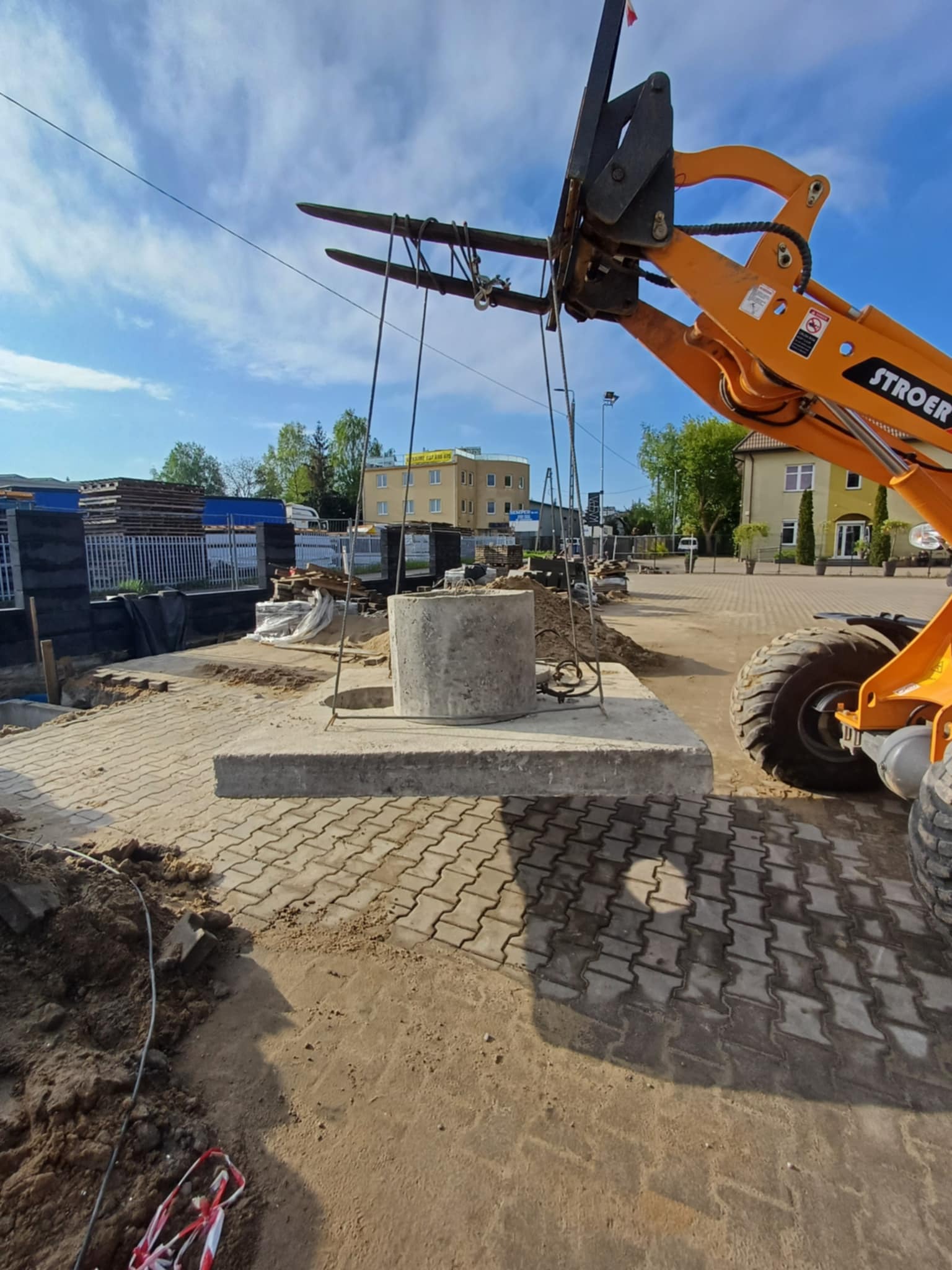 Pomarańczowy wózek widłowy transportuje betonowy element na placu budowy z kostką brukową w tle, widoczny budynek i drzewa.