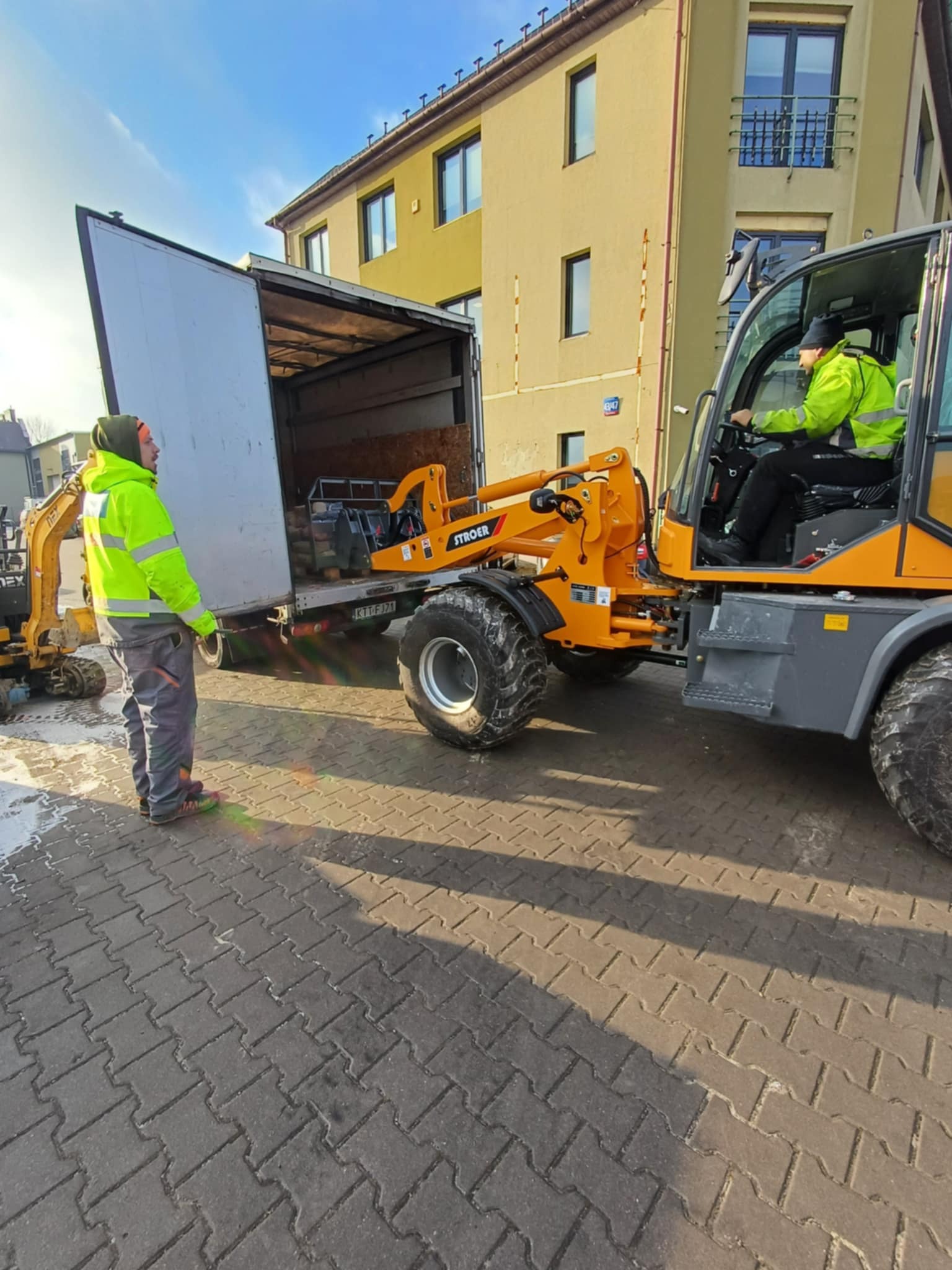 Rozładunek materiałów budowlanych z ciężarówki za pomocą pomarańczowej ładowarki teleskopowej na kostce brukowej, w tle budynek i pracownik w żółtej kamizelce.