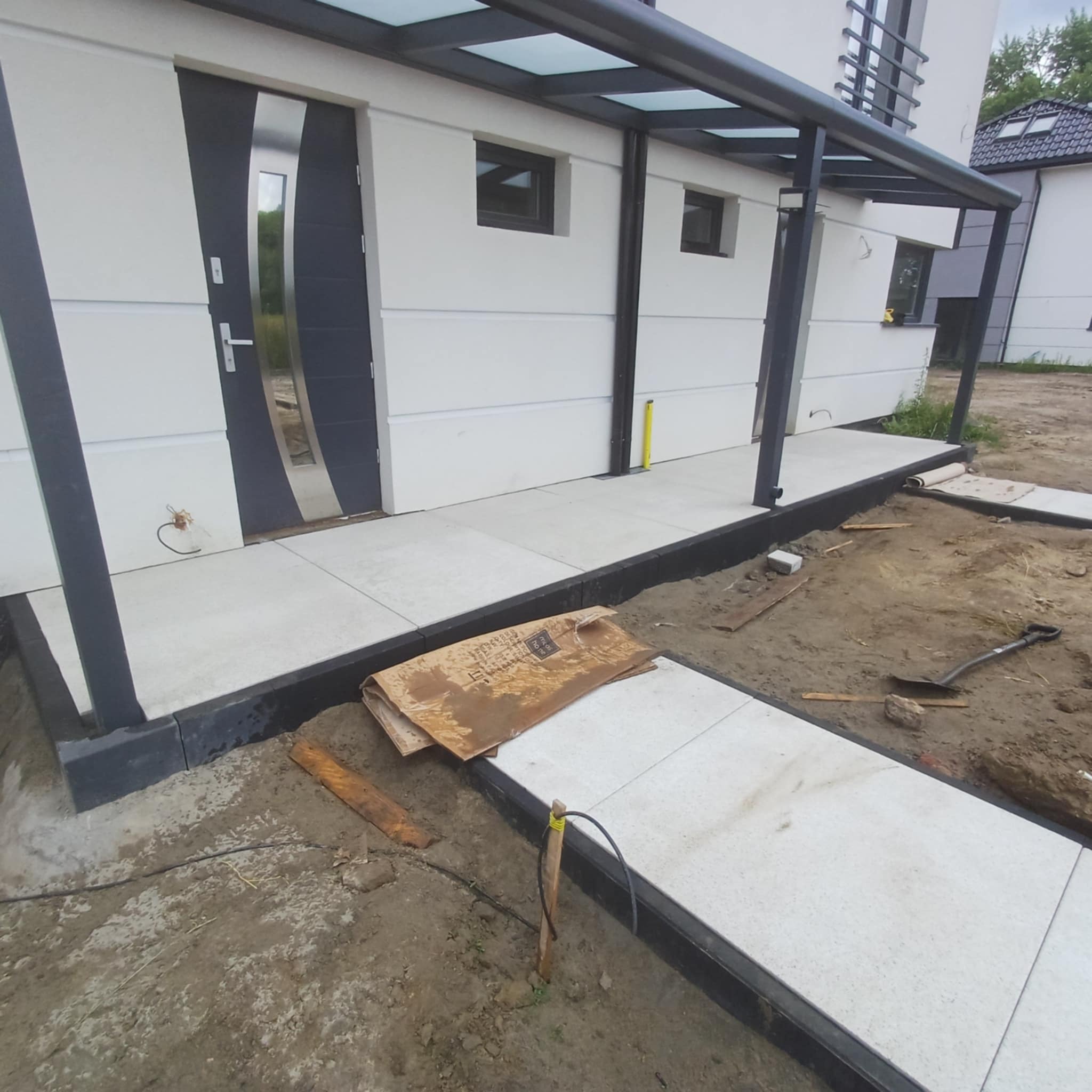Układanie jasnej kostki brukowej przed wejściem do nowoczesnego domu. Widoczny fragment budynku z zadaszeniem, ziemia, narzędzia. Częściowo ukończony chodnik.