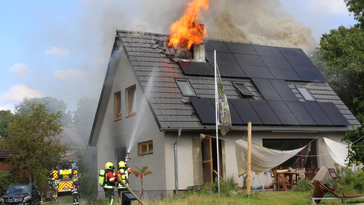 Płonący dom z panelami słonecznymi na dachu, gaszony przez strażaków. Widoczny wóz strażacki i dym. Ogień wydobywa się z dachu w okolicy komina.
