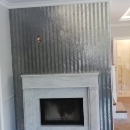 MAD-DOM Madejczyk Kacper - Elegancki kominek z marmurową obudową i dekoracyjną ścianą z błyszczących, srebrnych płytek ułożonych we wzór jodełki. Widoczne detale wykończeniowe i oświetlenie.