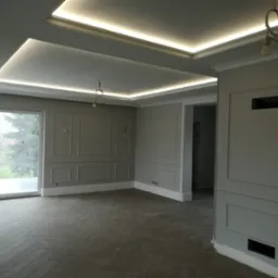 Eleganckie wykończenie wnętrza: szara kolorystyka, dekoracyjne listwy ścienne, parkiet w jodełkę i podświetlany sufit. Widok na okno i fragment instalacji elektrycznej.