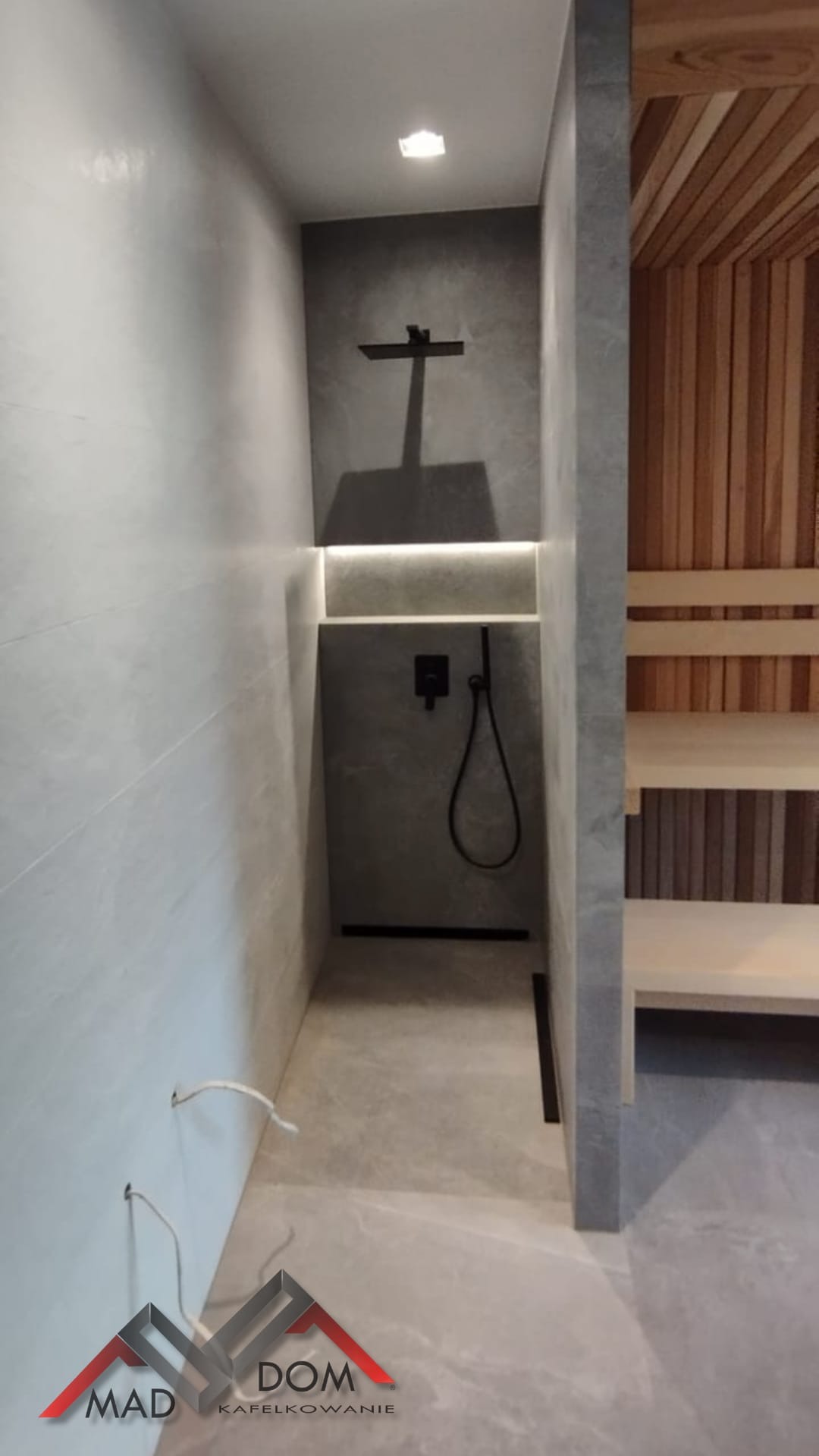Nowoczesna łazienka z kabiną prysznicową typu walk-in, wykończona w szarościach, z elementami drewna. Widoczne minimalistyczne baterie i oświetlenie LED.