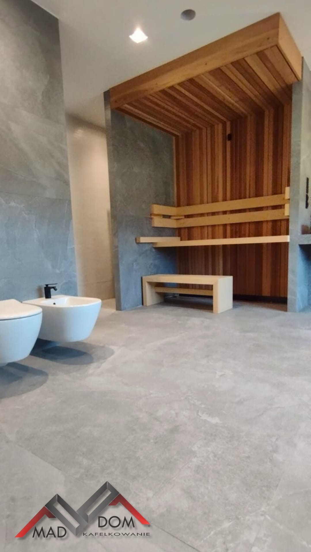 Nowoczesna łazienka z sauną: szare płytki imitujące kamień, drewniana sauna z siedziskami, białe sanitariaty. Minimalistyczny design i naturalne materiały.