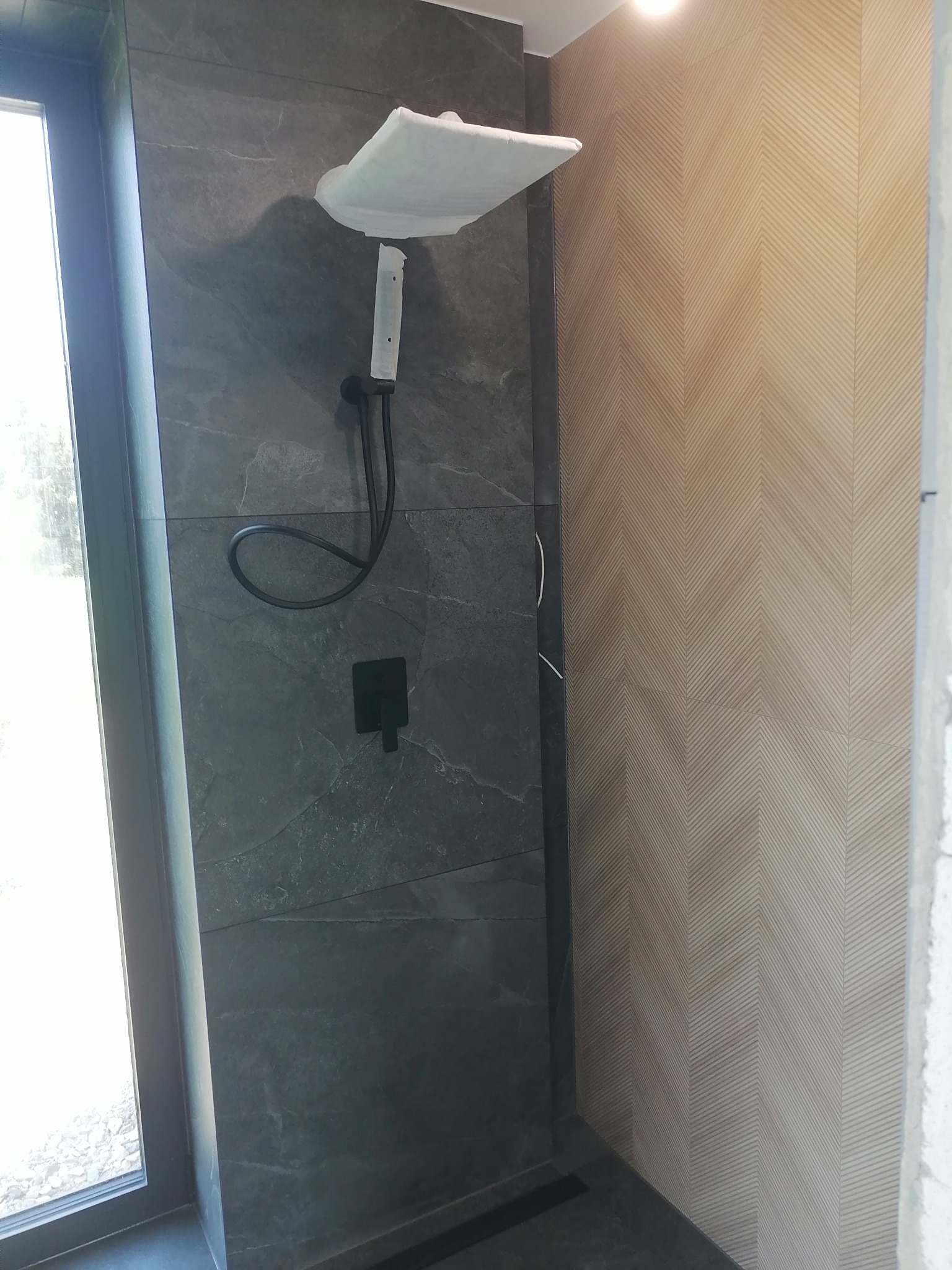 Nowoczesna kabina prysznicowa z grafitowymi płytkami imitującymi kamień i beżową ścianą w geometryczny wzór, jeszcze zabezpieczona folią ochronną.