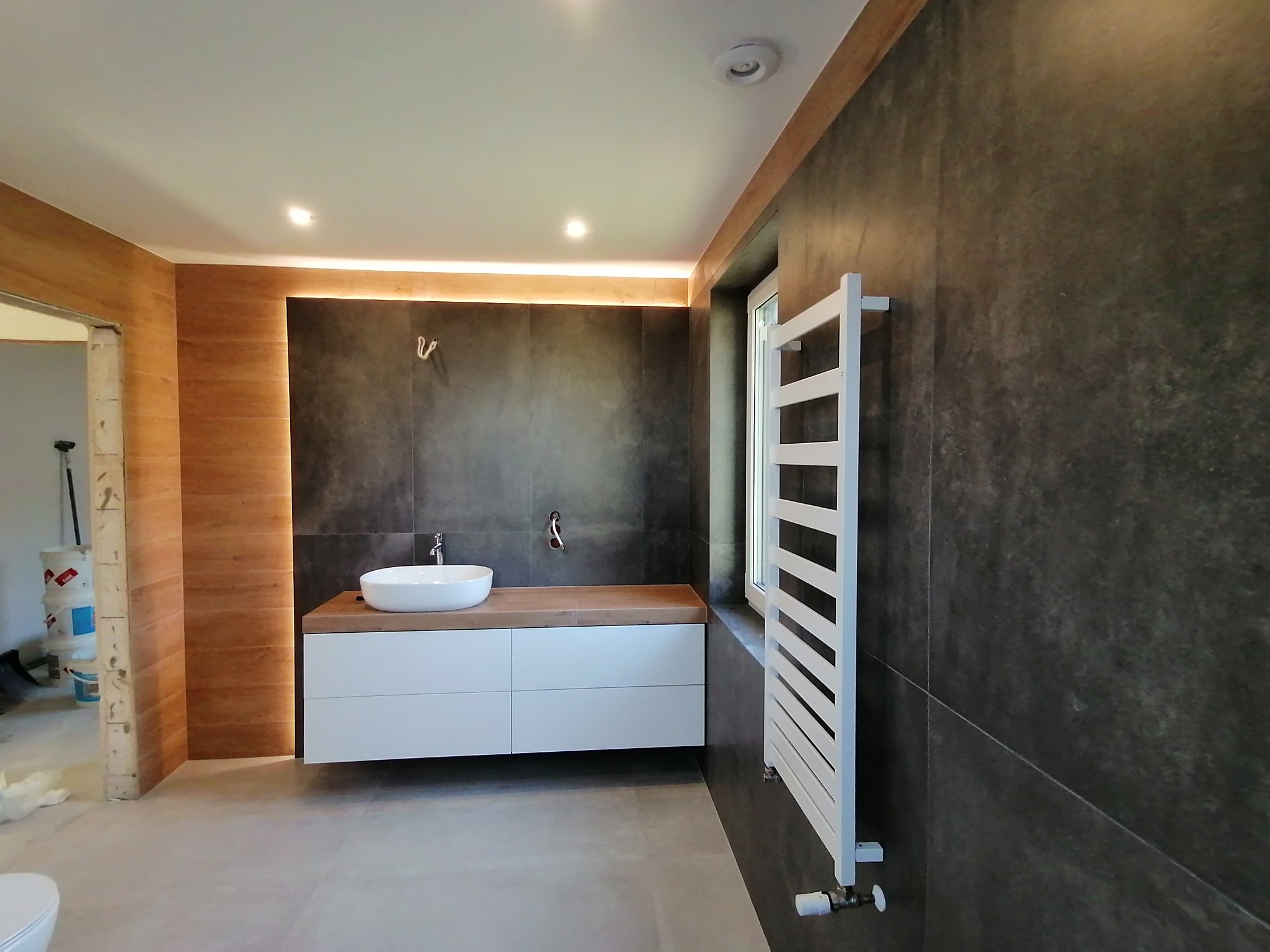 Nowoczesna łazienka z umywalką nablatową, grzejnikiem drabinkowym i drewnianymi akcentami. Widoczny fragment nieukończonego przejścia.