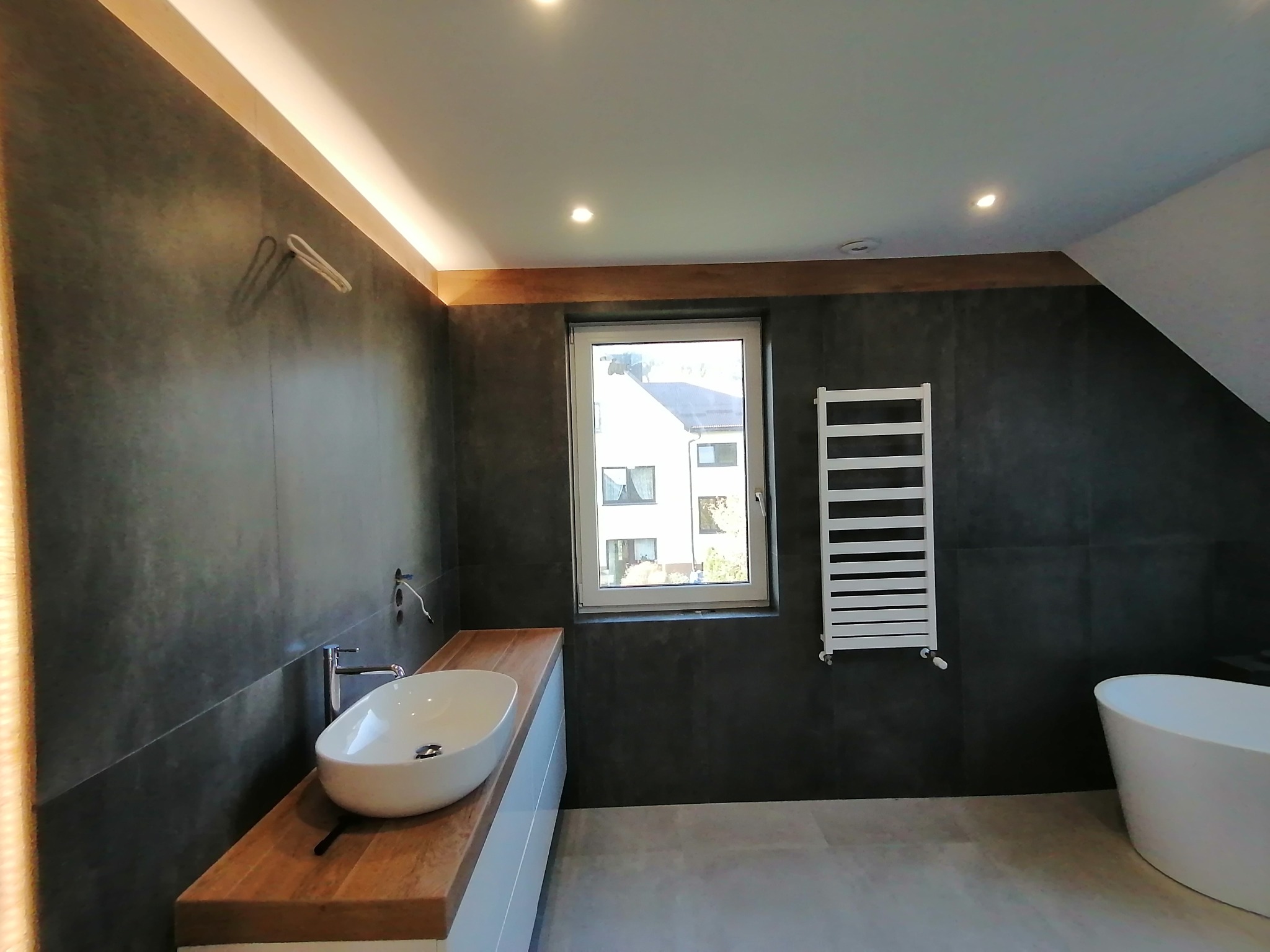 Nowoczesna łazienka z szarymi płytkami, drewnianym blatem pod umywalką i białą grzejnikiem drabinkowym. Widoczne okno i wanna wolnostojąca.