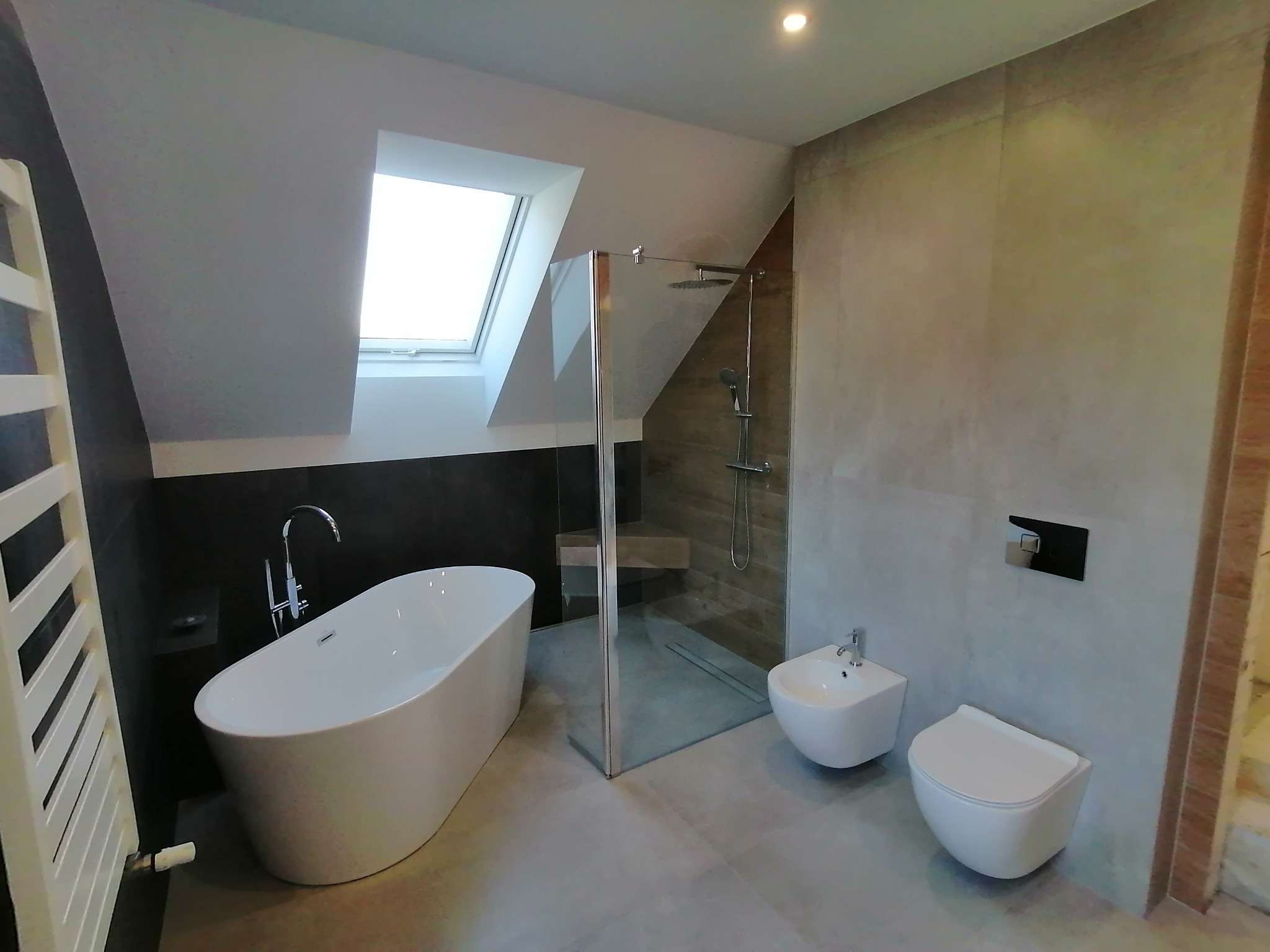 Nowoczesna łazienka na poddaszu: wolnostojąca wanna, kabina prysznicowa ze szklaną ścianą, bidet i toaleta. Ściany wykończone w szarościach, okno dachowe.