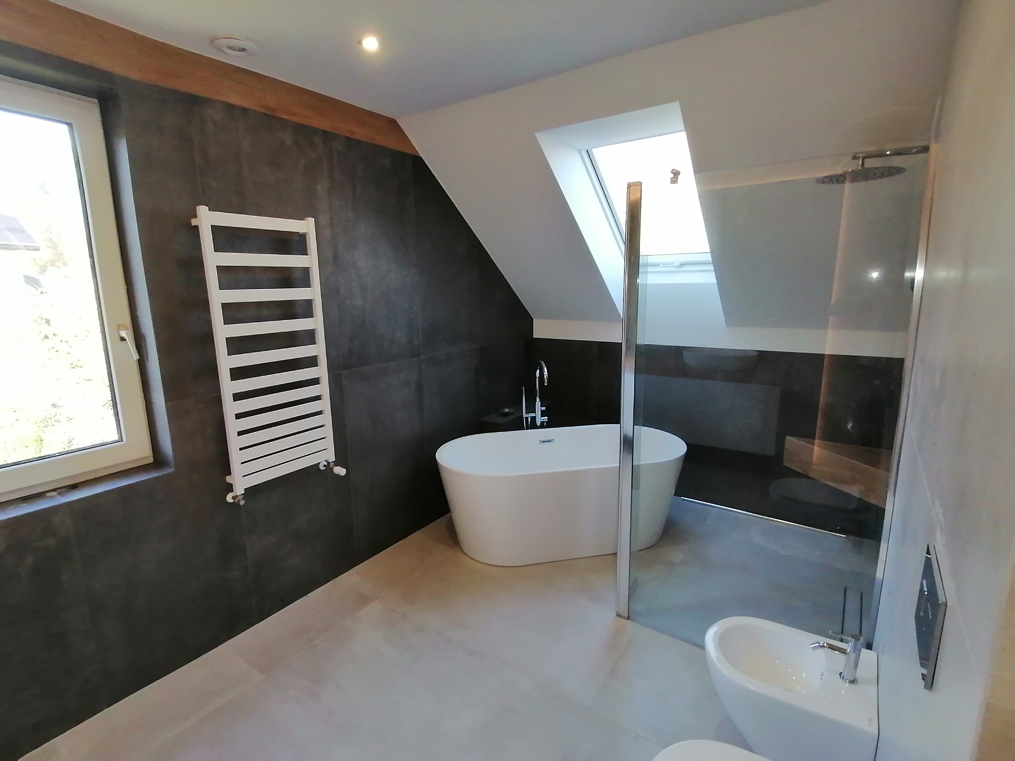 Nowoczesna łazienka z wolnostojącą wanną, grzejnikiem drabinkowym i kabiną prysznicową ze szklaną ścianką. Uwagę zwraca okno dachowe i ciemne płytki.