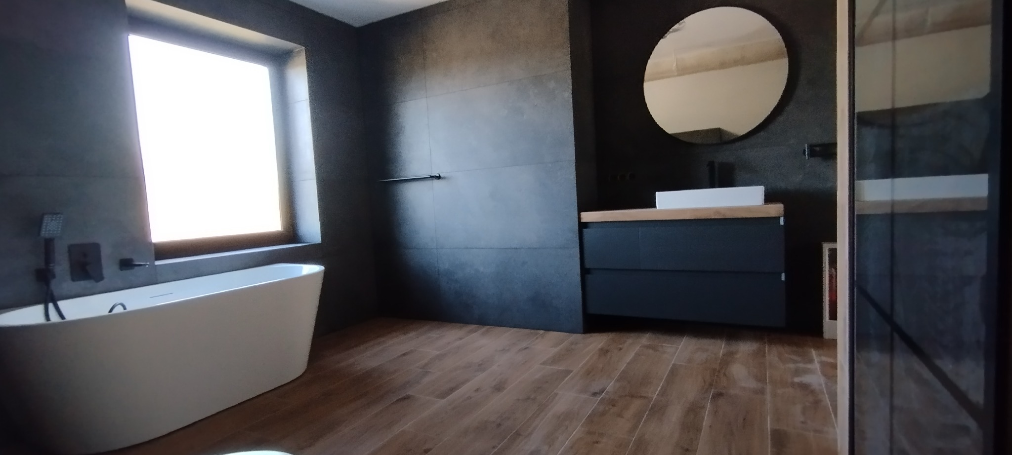 Nowoczesna łazienka z drewnianą podłogą, ciemnymi płytkami i owalnym lustrem. Biała wanna wolnostojąca pod oknem. Minimalistyczny design.