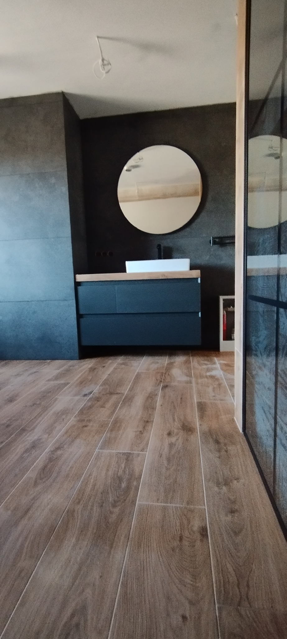 Nowoczesna łazienka z drewnopodobną podłogą, grafitowymi ścianami i okrągłym lustrem nad umywalką nablatową. Kabina prysznicowa z czarnymi szprosami.