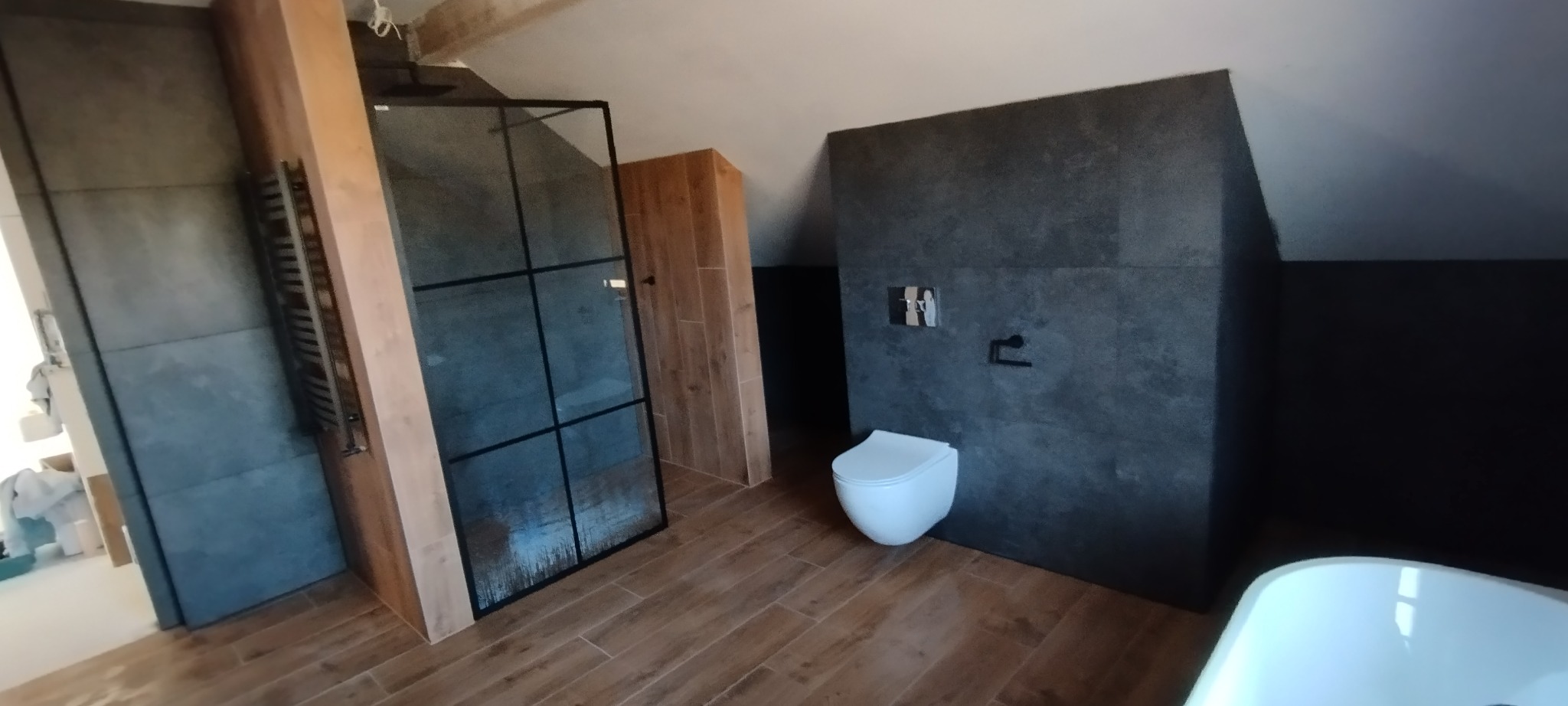 Nowoczesna łazienka na poddaszu ze szklaną kabiną, drewnianymi akcentami i szarą ścianą z wiszącą toaletą. Podłoga wyłożona płytkami imitującymi drewno.