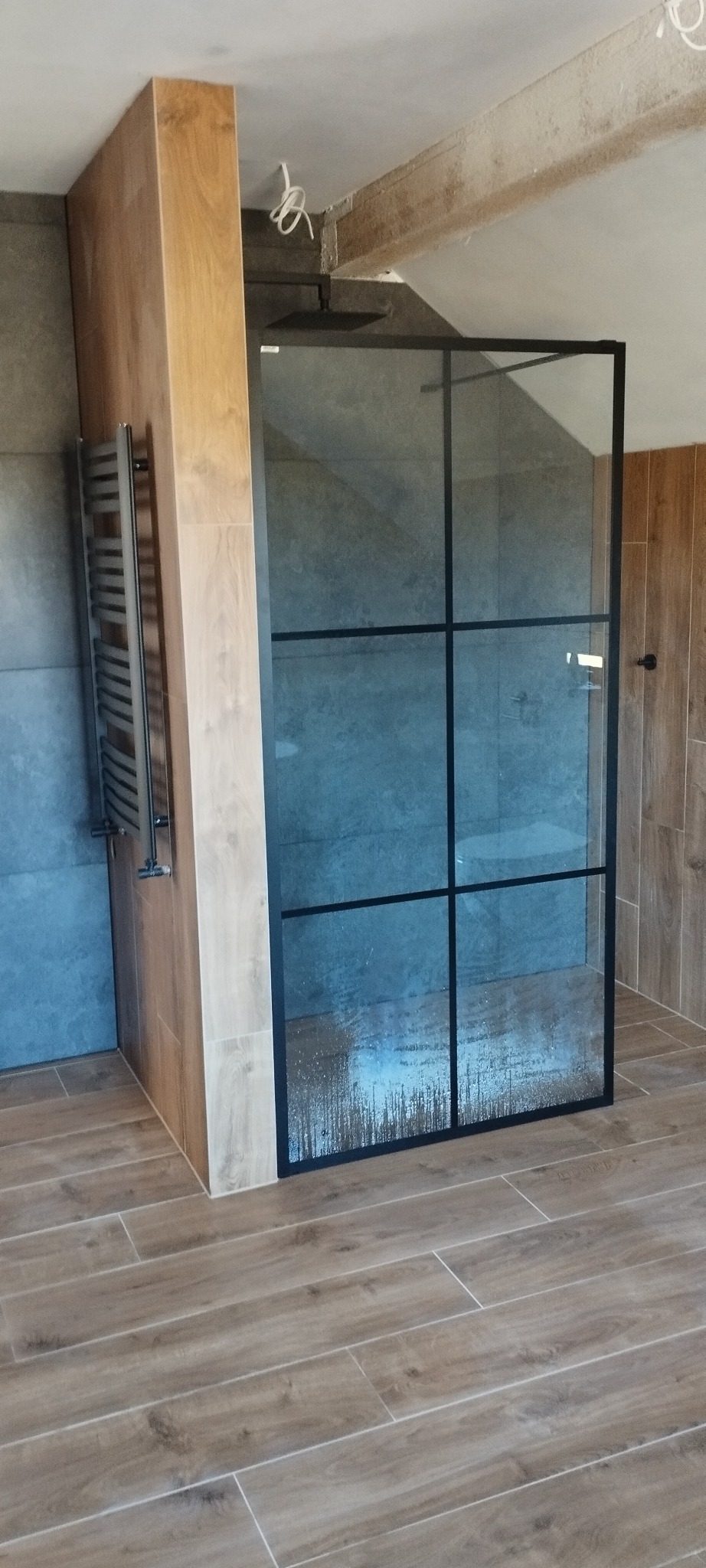 Szklana kabina prysznicowa z czarnymi szprosami w wykończonej łazience z płytkami imitującymi drewno, grzejnik drabinkowy po lewej stronie.