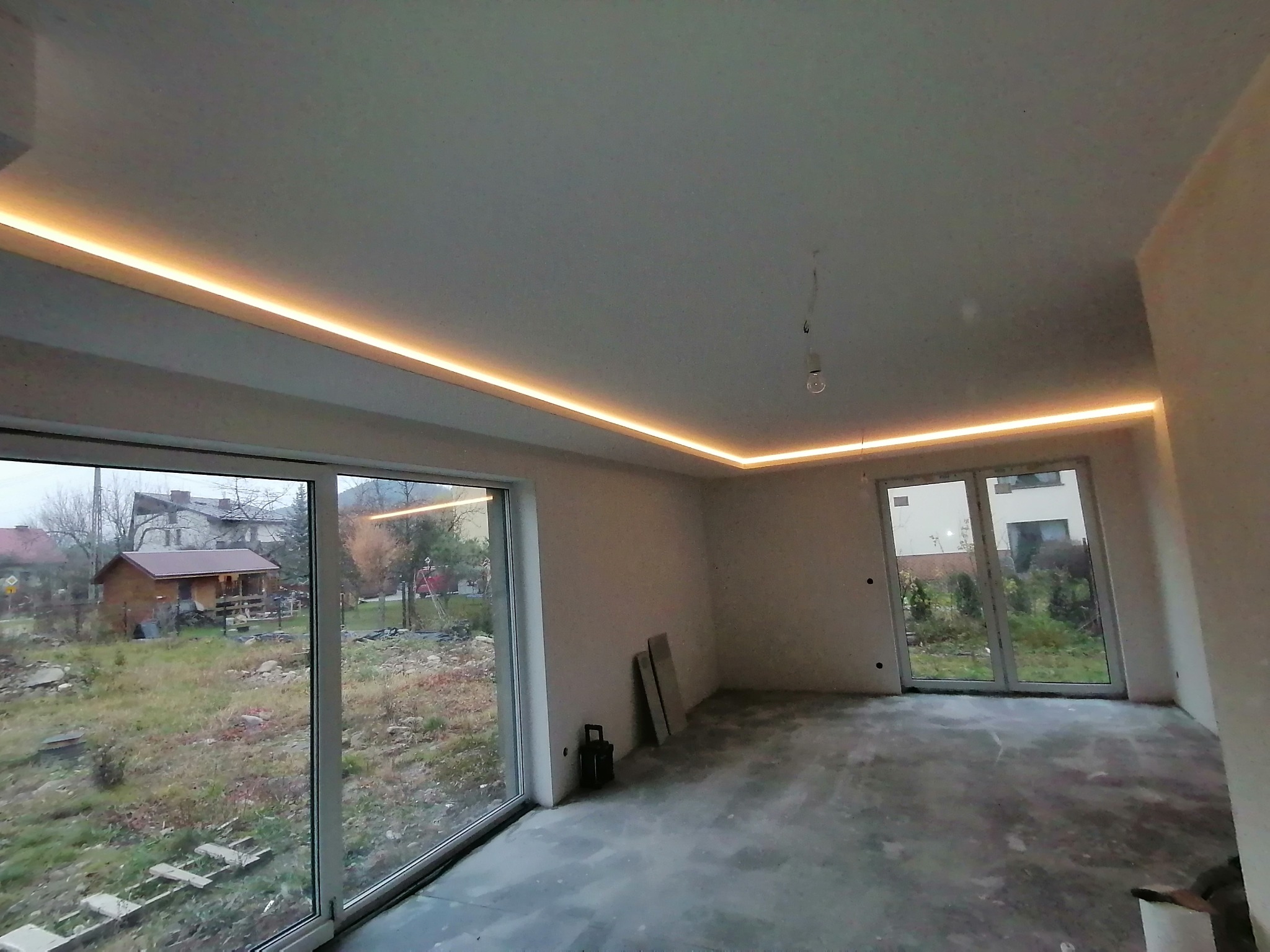 Wykończone wnętrze pokoju z oświetleniem LED na suficie, widoczne duże okna z widokiem na zewnątrz i surowa podłoga. Ściany przygotowane do malowania.