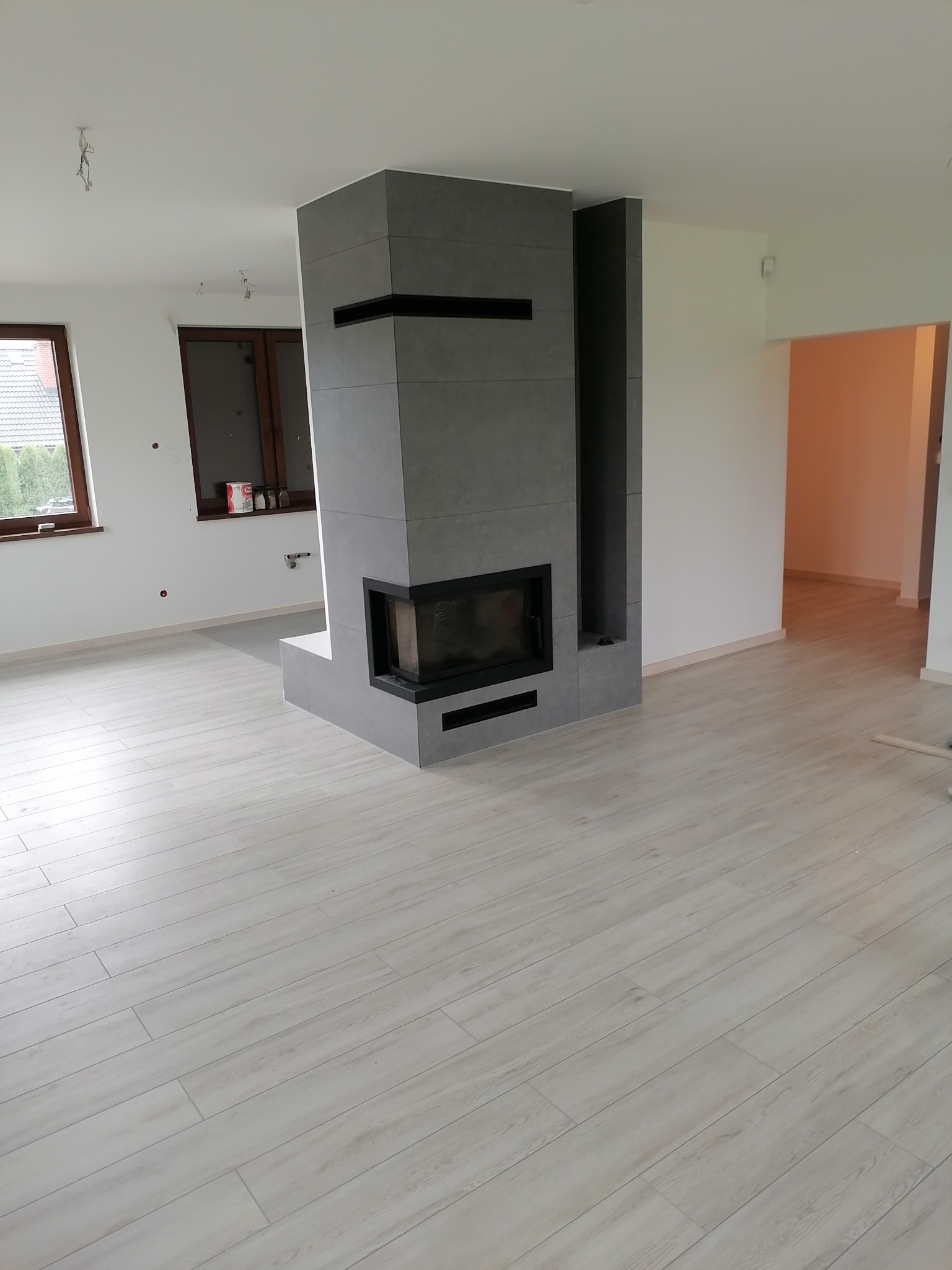 Nowoczesny salon z jasną podłogą i obudowanym kominkiem z szarych płyt. Widoczne okno i fragment niezakończonych prac wykończeniowych.