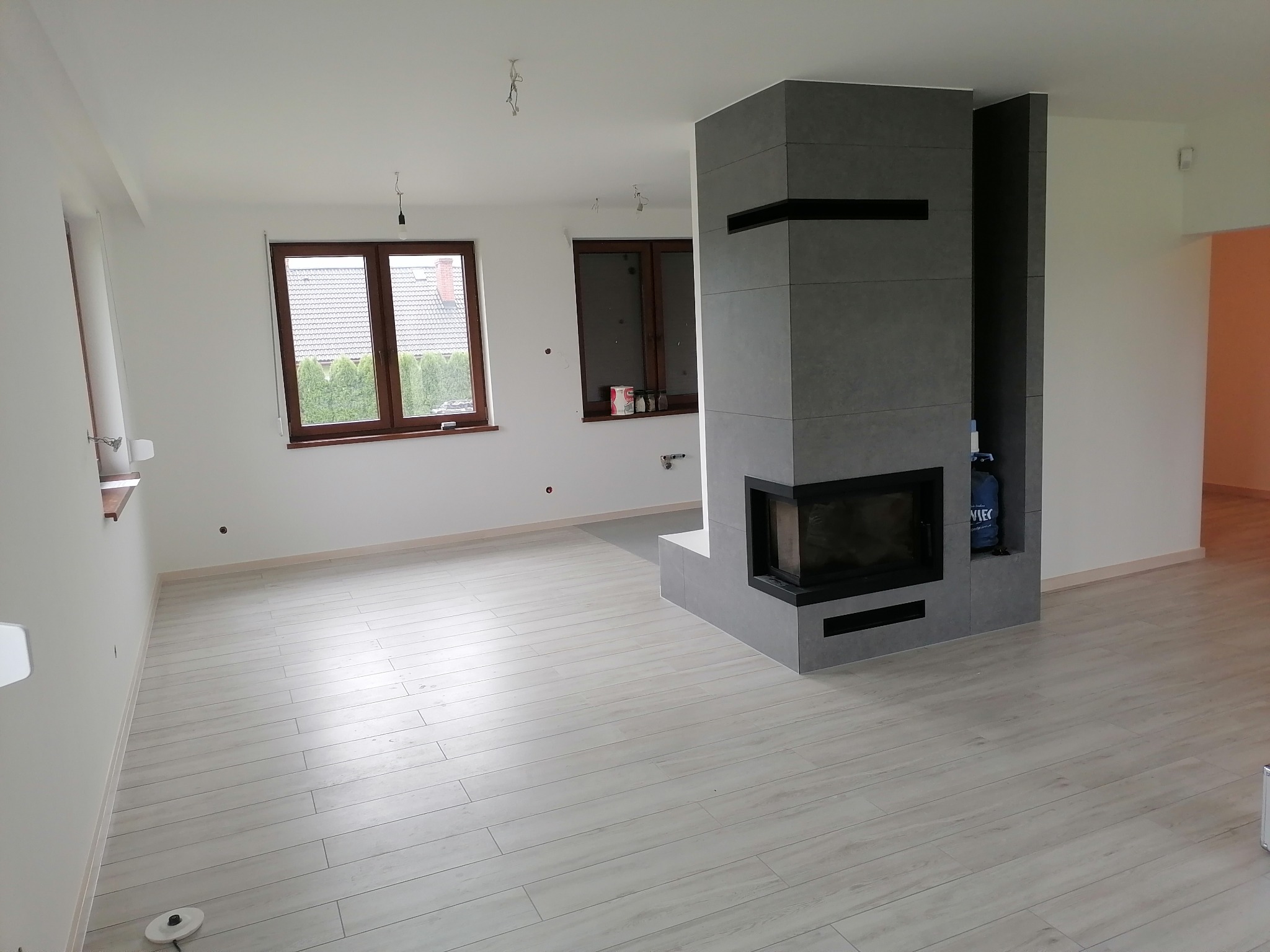 Jasne, przestronne wnętrze po remoncie z wykończonym kominkiem obłożonym szarą płytką i drewnopodobną podłogą. Widoczne dwa okna i niedokończone punkty oświetleniowe.