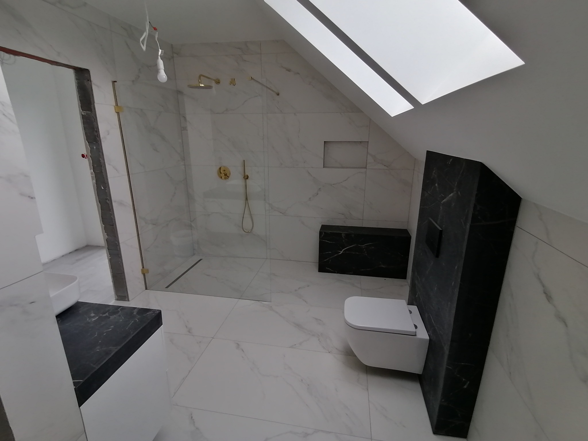 Wykończona łazienka na poddaszu z marmurowymi płytkami, kabiną prysznicową ze złotą armaturą i wiszącą toaletą obudowaną czarnym marmurem. Widoczne okno dachowe.