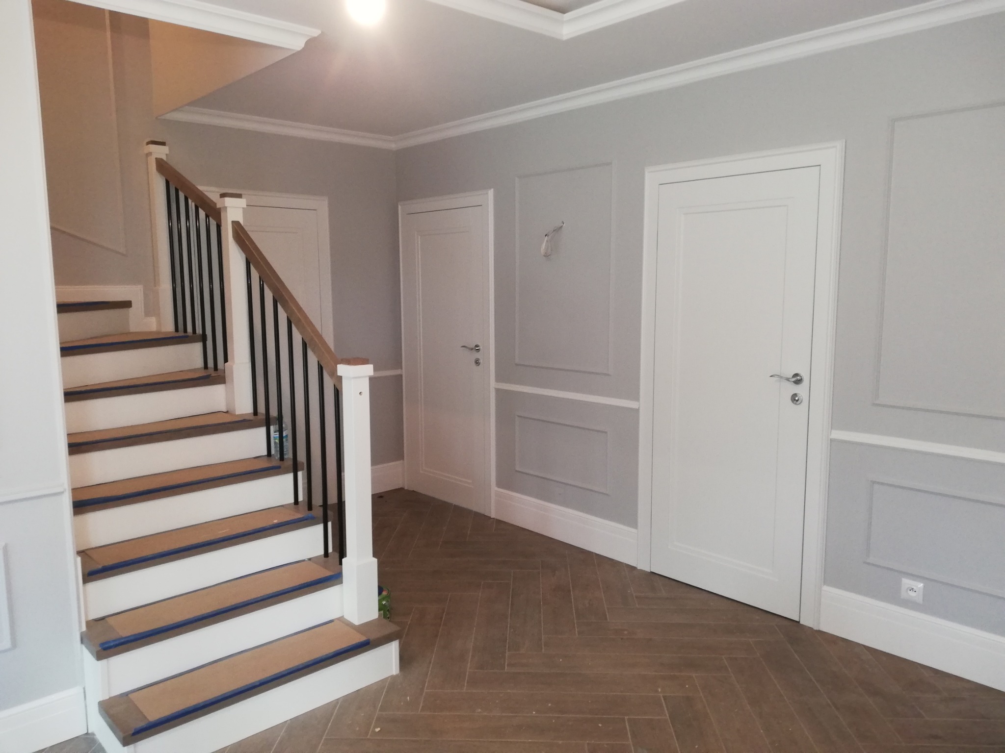 Korytarz po remoncie: szare ściany z dekoracyjnymi panelami, białe drzwi, drewniana podłoga w jodełkę i schody z czarną balustradą. Widoczne listwy przypodłogowe.