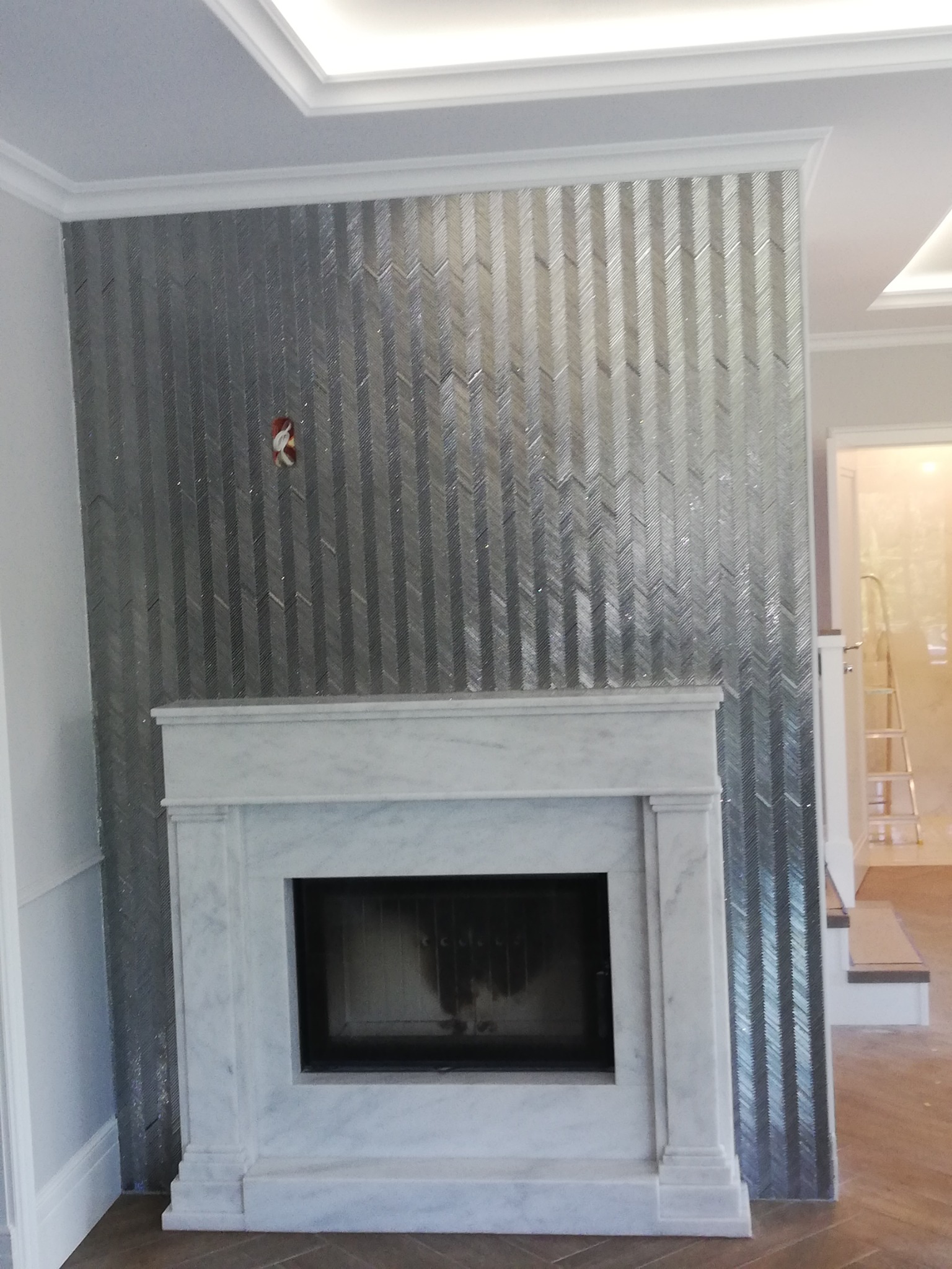 Elegancki kominek z marmurową obudową i dekoracyjną ścianą z błyszczących, srebrnych płytek ułożonych we wzór jodełki. Widoczne detale wykończeniowe i oświetlenie.