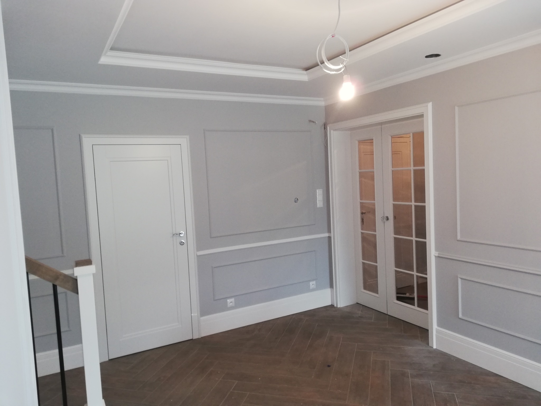 Eleganckie wykończenie wnętrza w szarościach: panele ścienne z listwami, białe drzwi i parkiet w jodełkę. Surowe oświetlenie podkreśla detale.