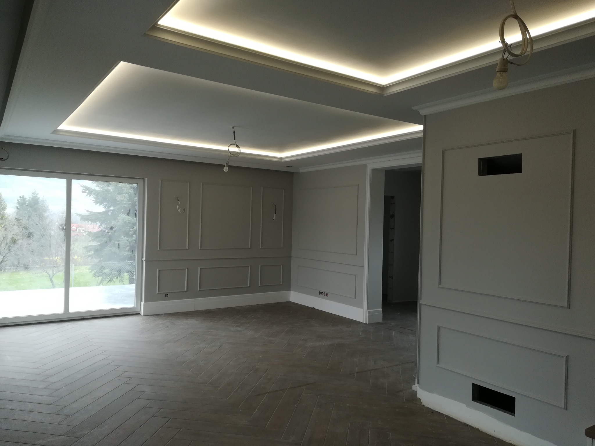 Eleganckie wykończenie wnętrza: szara kolorystyka, dekoracyjne listwy ścienne, parkiet w jodełkę i podświetlany sufit. Widok na okno i fragment instalacji elektrycznej.