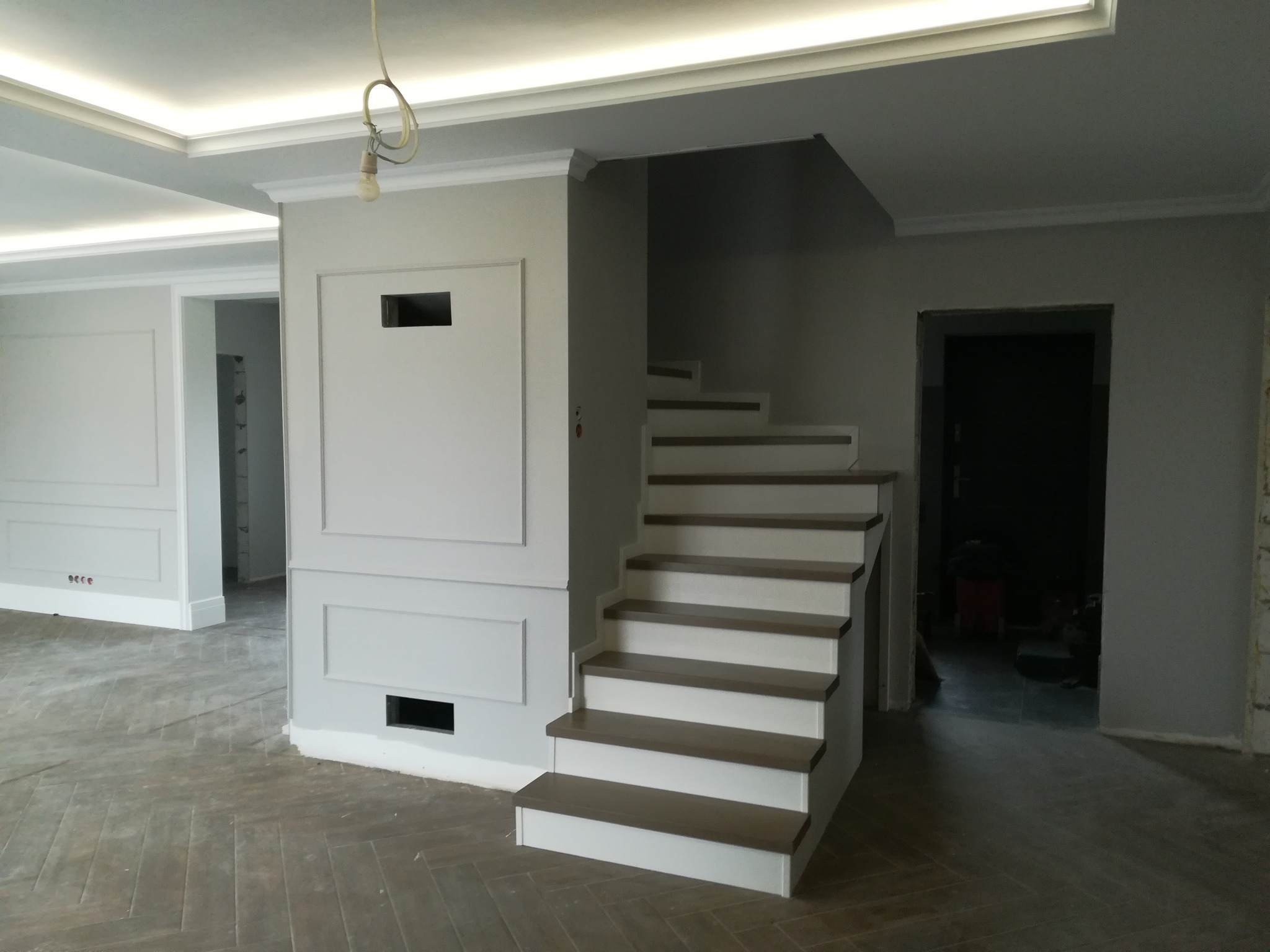 Jasne wnętrze z dekoracyjnymi panelami ściennymi, schodami z drewnianymi stopniami i oświetleniem sufitowym. Podłoga wyłożona parkietem w jodełkę.