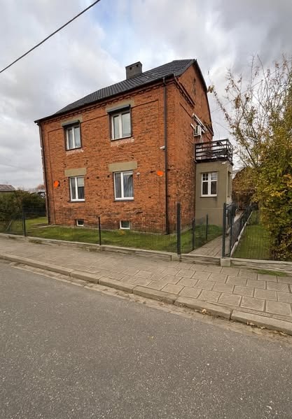 Dwupiętrowy dom z czerwonej cegły z balkonem, widoczny z poziomu ulicy w pochmurny dzień. Nowe okna z roletami i czarny dach z kominem. Metalowe ogrodzenie.