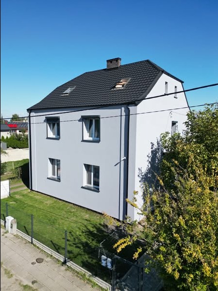 Dwukondygnacyjny dom z szarą elewacją i ciemnym dachem, widziany z góry pod kątem. Widoczne okna, komin i fragment zieleni. Słoneczny dzień, błękitne niebo.