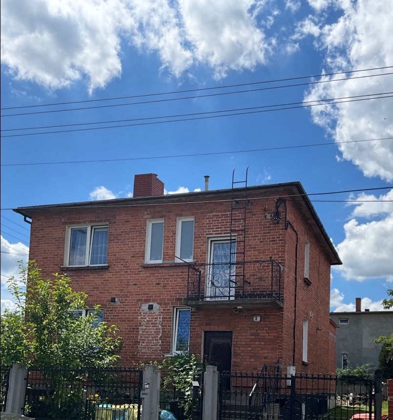 Dwupiętrowy dom z czerwonej cegły z balkonem i drabinką na dachu, otoczony ogrodzeniem. Część elewacji z śladami po pracach. Błękitne niebo z białymi chmurami w tle.