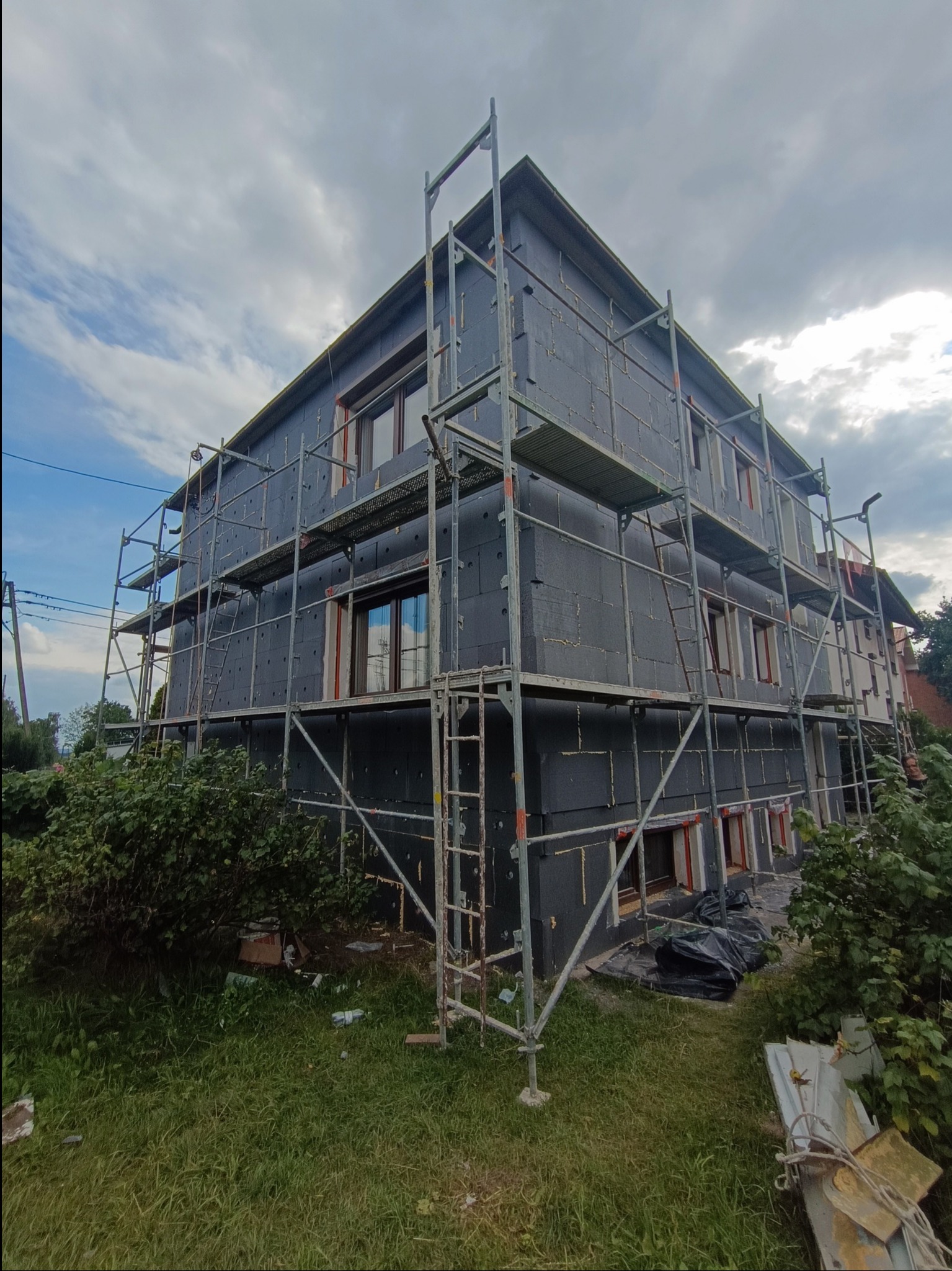 Dwukondygnacyjny dom w trakcie ocieplania elewacji styropianem, otoczony rusztowaniem. Widoczne detale: czarny styropian, okna z czerwonymi ramami, zielona trawa i krzewy.