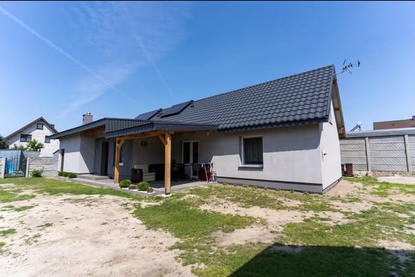 Dom jednorodzinny z panelami słonecznymi na dachu i drewnianą pergolą, widok z zewnątrz w słoneczny dzień. Szara elewacja i dach z ciemnej dachówki.