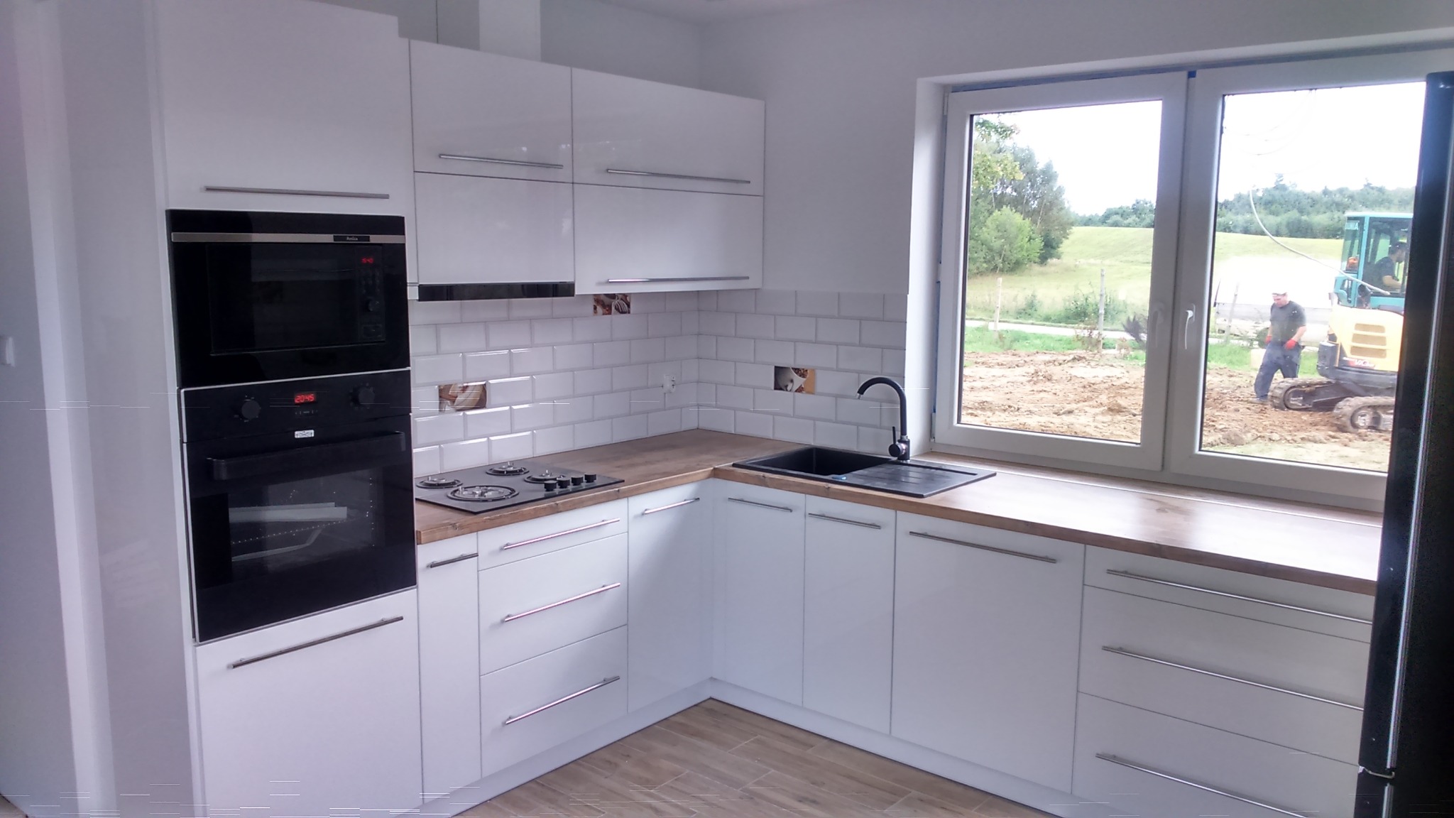 Biała, narożna zabudowa kuchenna z drewnianym blatem i płytkami imitującymi cegłę. W tle widok z okna na prace ziemne z użyciem koparki.
