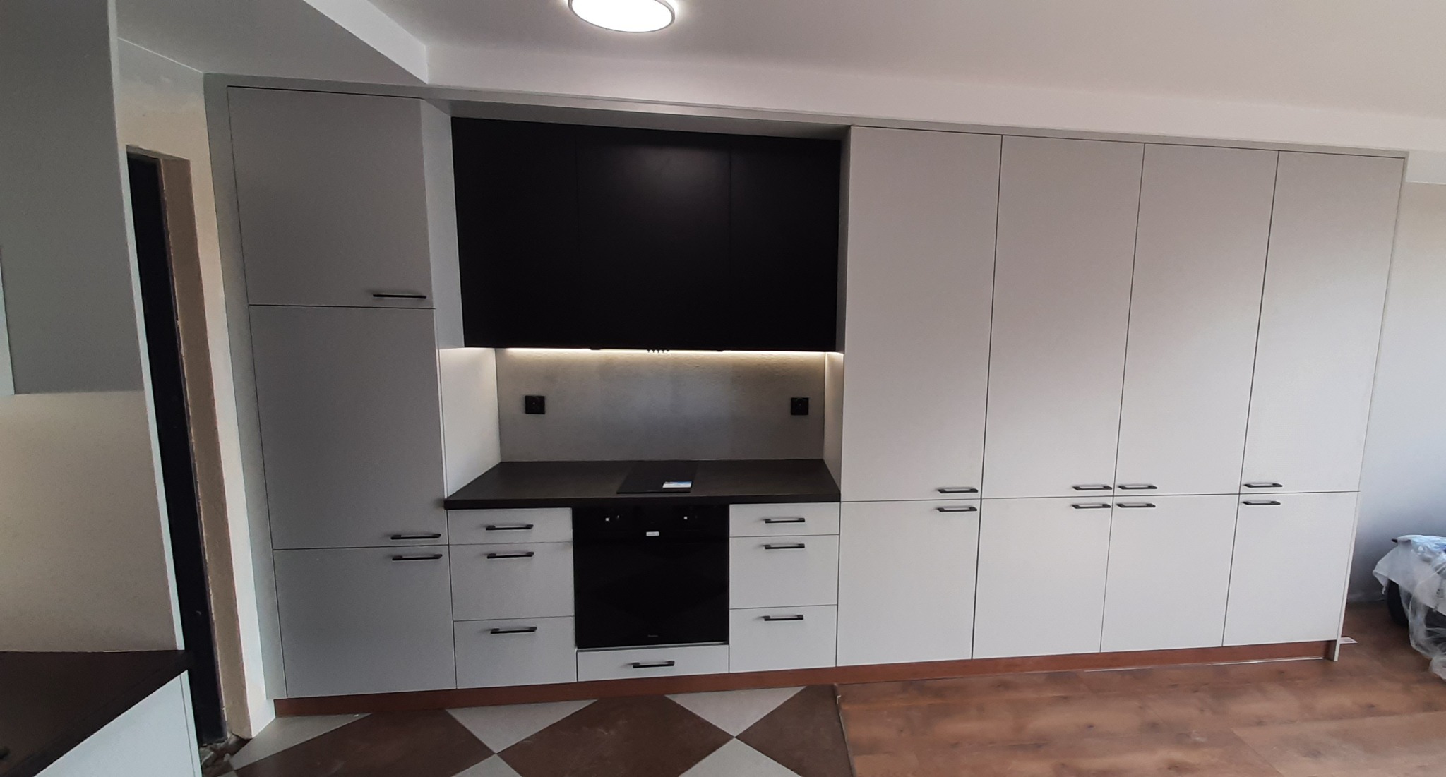 Nowoczesna kuchnia na wymiar z szarymi frontami i czarnymi szafkami wiszącymi, oświetleniem LED i brązową podłogą w szachownicę.